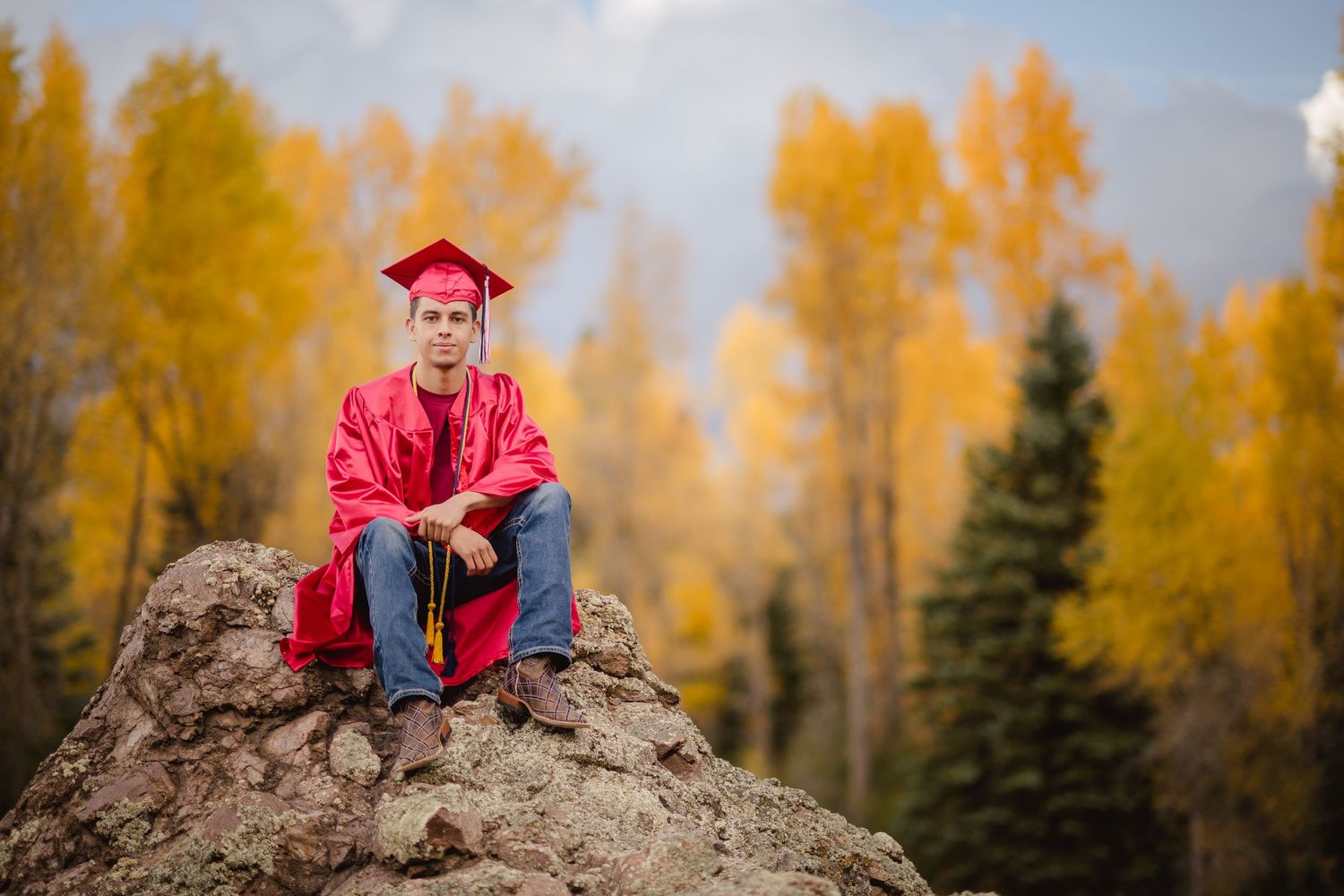 A graduate in red regalia sits on a rocky outcrop surrounded by golden autumn trees in a mountain landscape.
