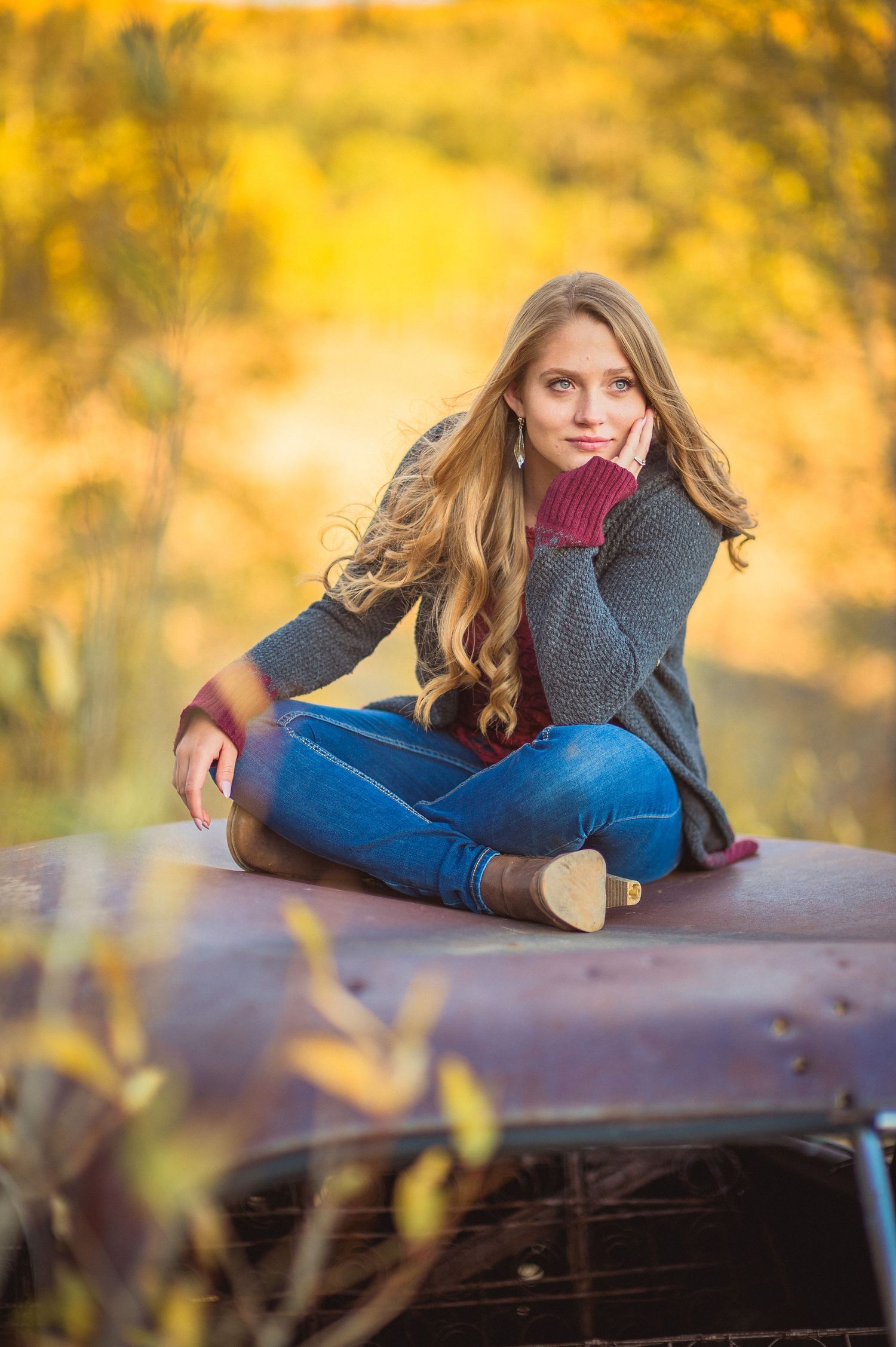 A person in a burgundy sweater and jeans lays on an outdoor surface with golden autumn foliage in the background.