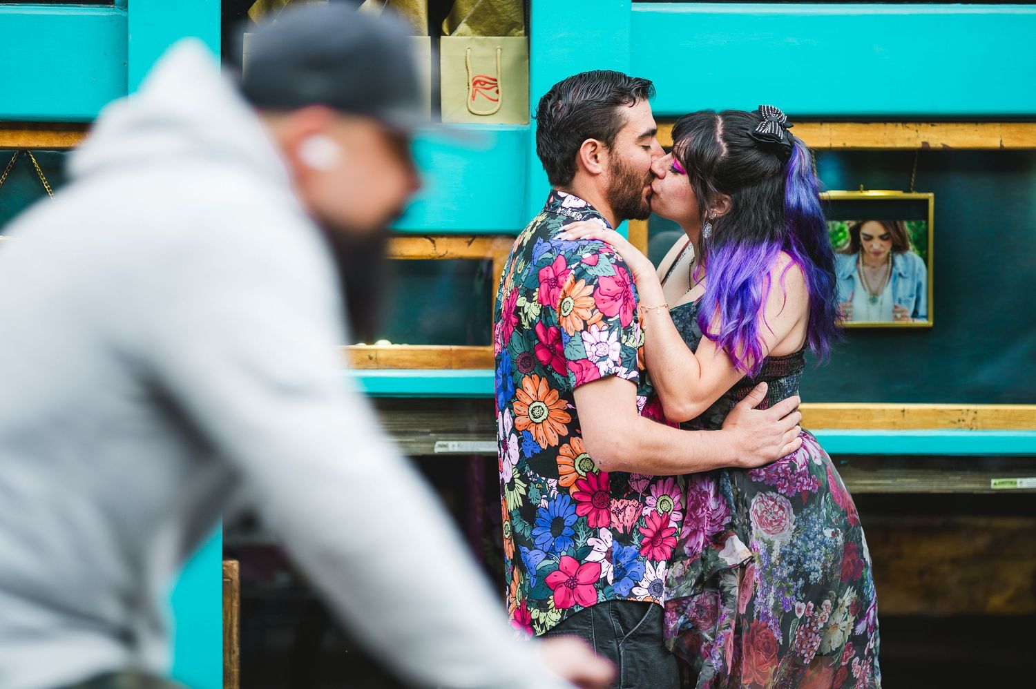 Couple kissing and embracing outdoors near a teal food truck, man in floral shirt, woman with purple hair.