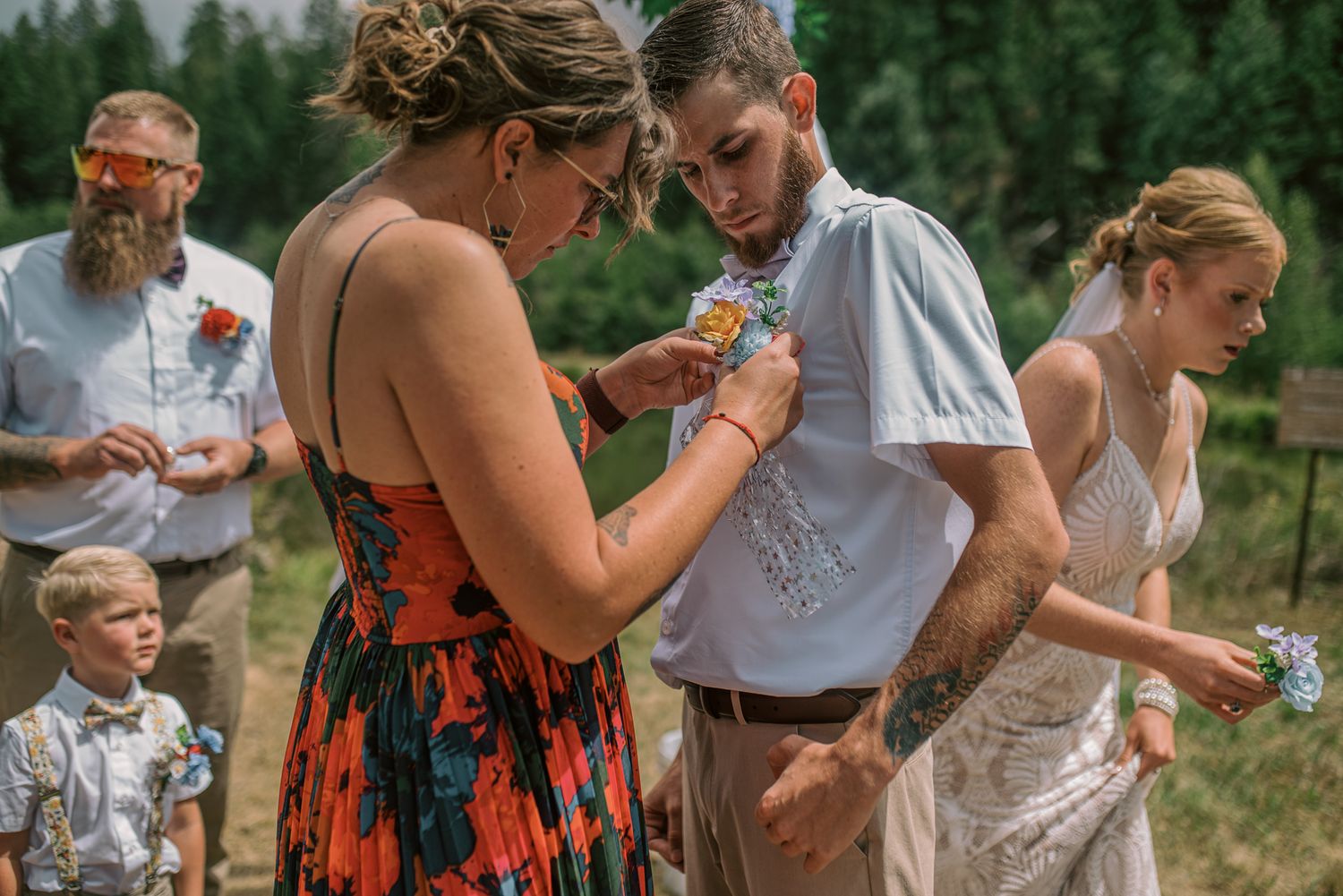 Wedding party pins boutonnieres on groom outdoors at rustic mountain ceremony with bride and guests.