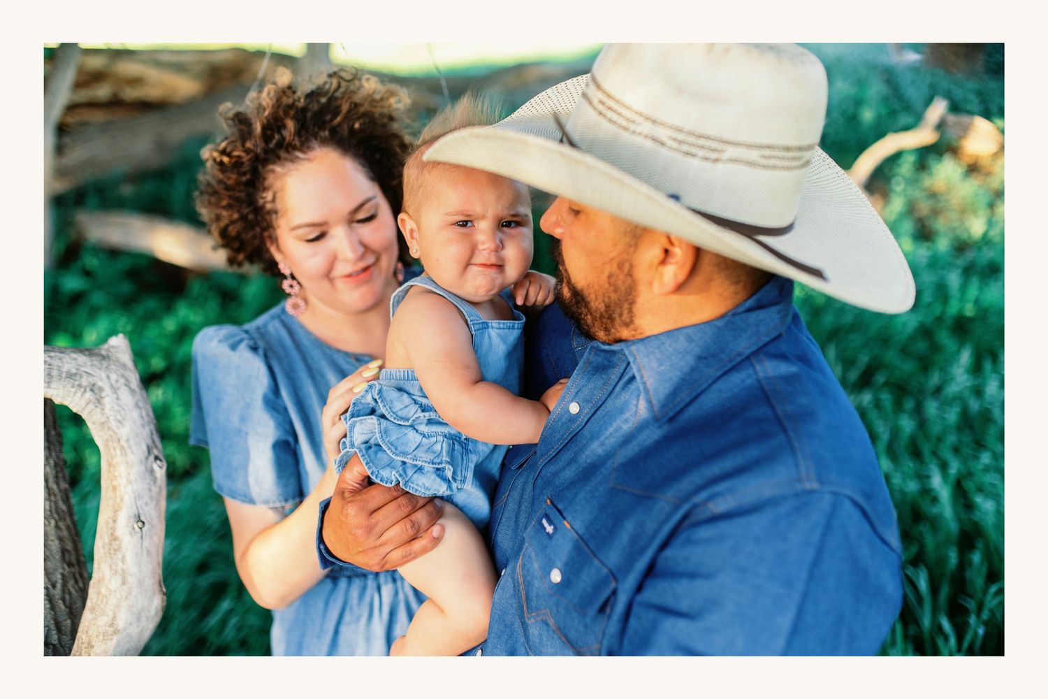 A couple in blue outfits holding their baby outdoors in a lush green setting, with the man wearing a cowboy hat.