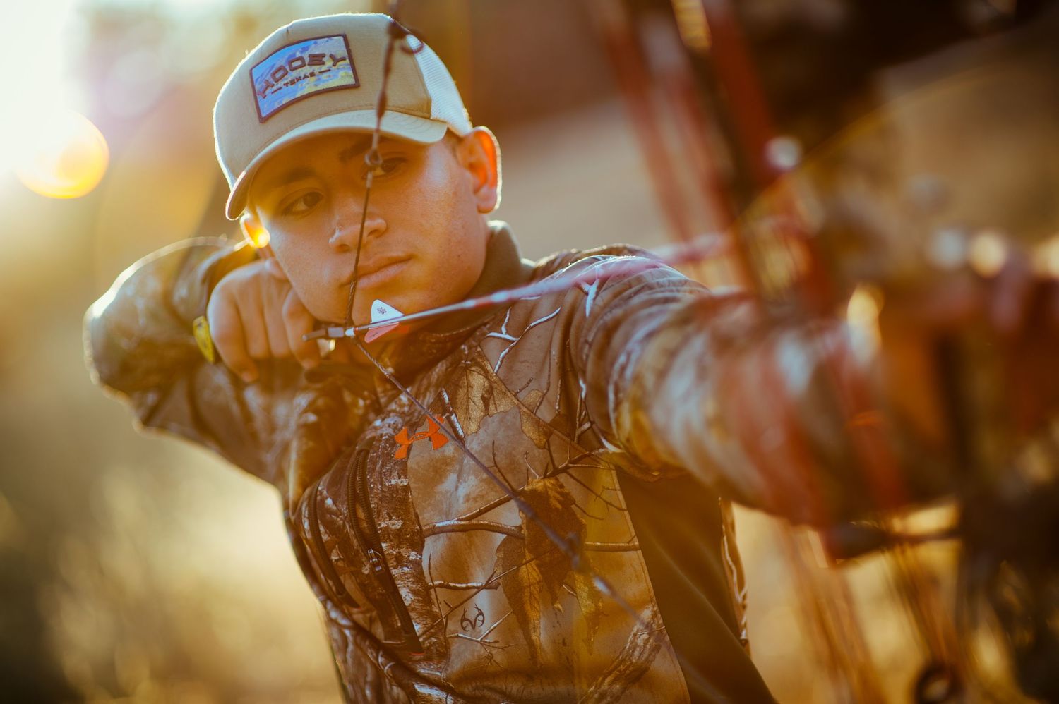 Young archer in camo jacket and trucker hat drawing a recurve bow outdoors in golden light.