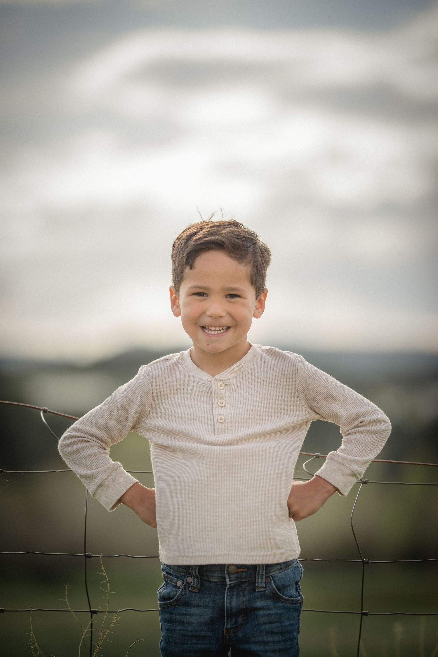 A young child in a cream henley shirt and jeans poses outdoors against a cloudy sky and wire fence backdrop.
