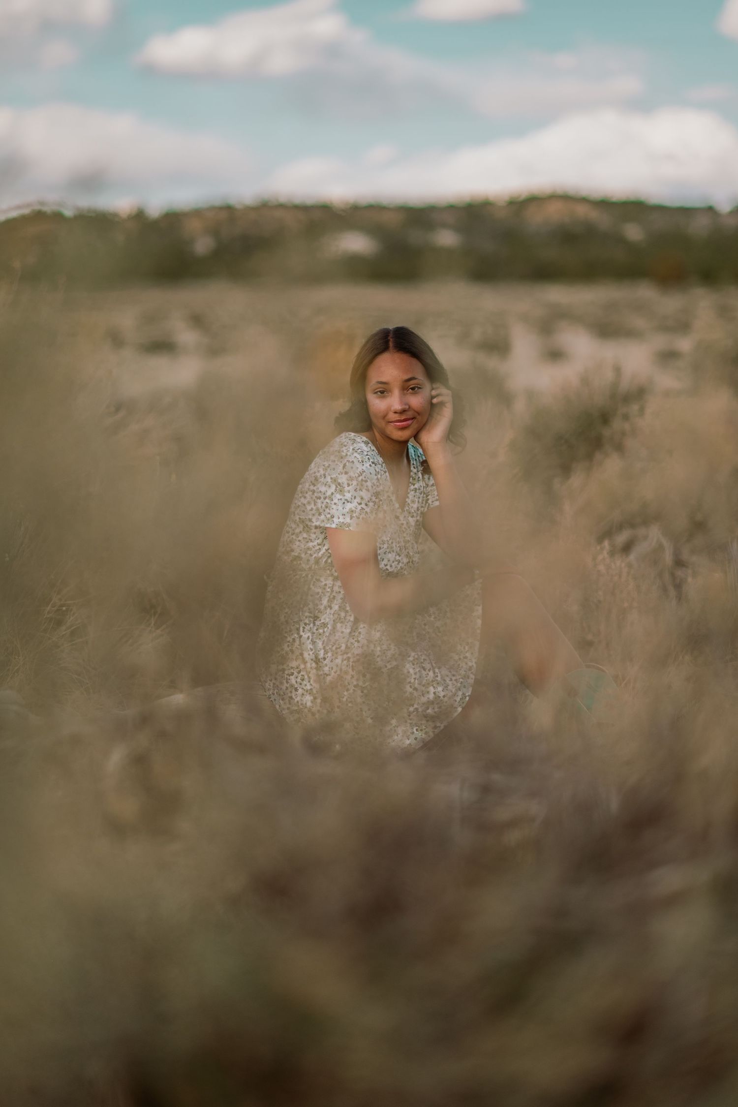 A person wearing a floral dress poses in a desert landscape with blurred brush and soft lighting in multiple shots.