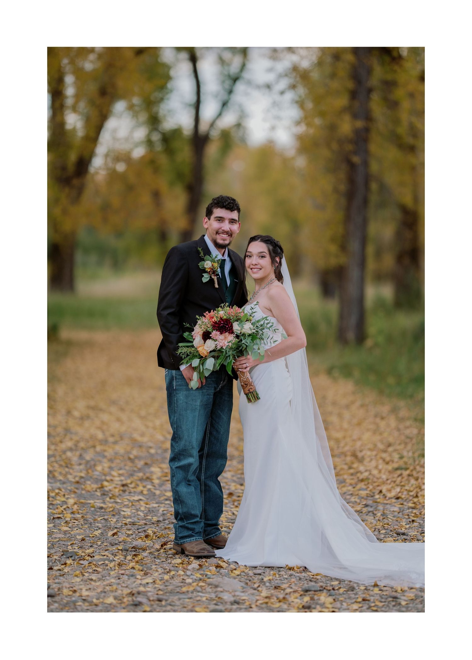 A couple poses together on a fall day surrounded by autumn leaves and trees, with the bride holding a colorful bouquet.