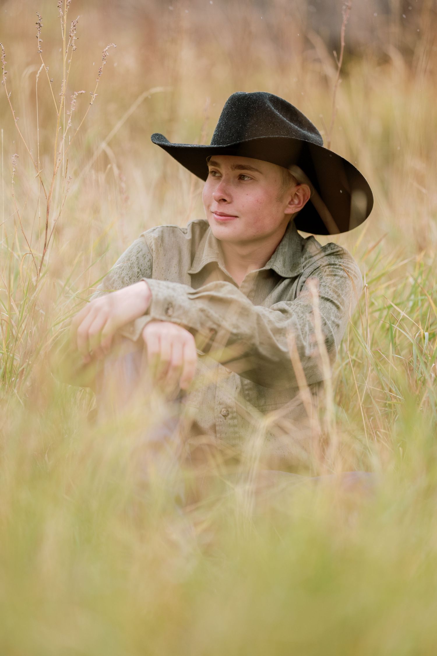 A cowboy in western attire sits contemplatively in tall prairie grass during a golden hour photoshoot.