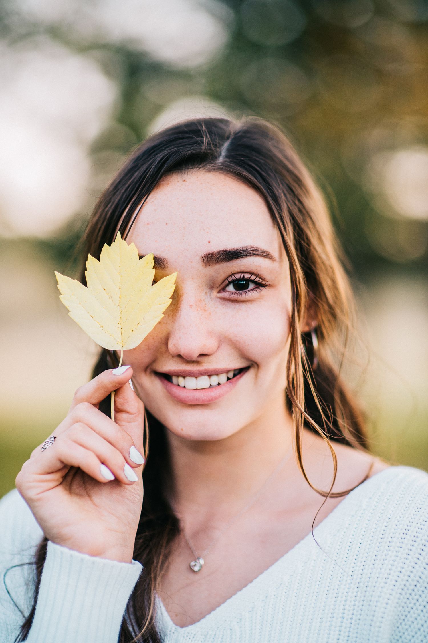 Person holding yellow autumn leaf near face while wearing white sweater outdoors.