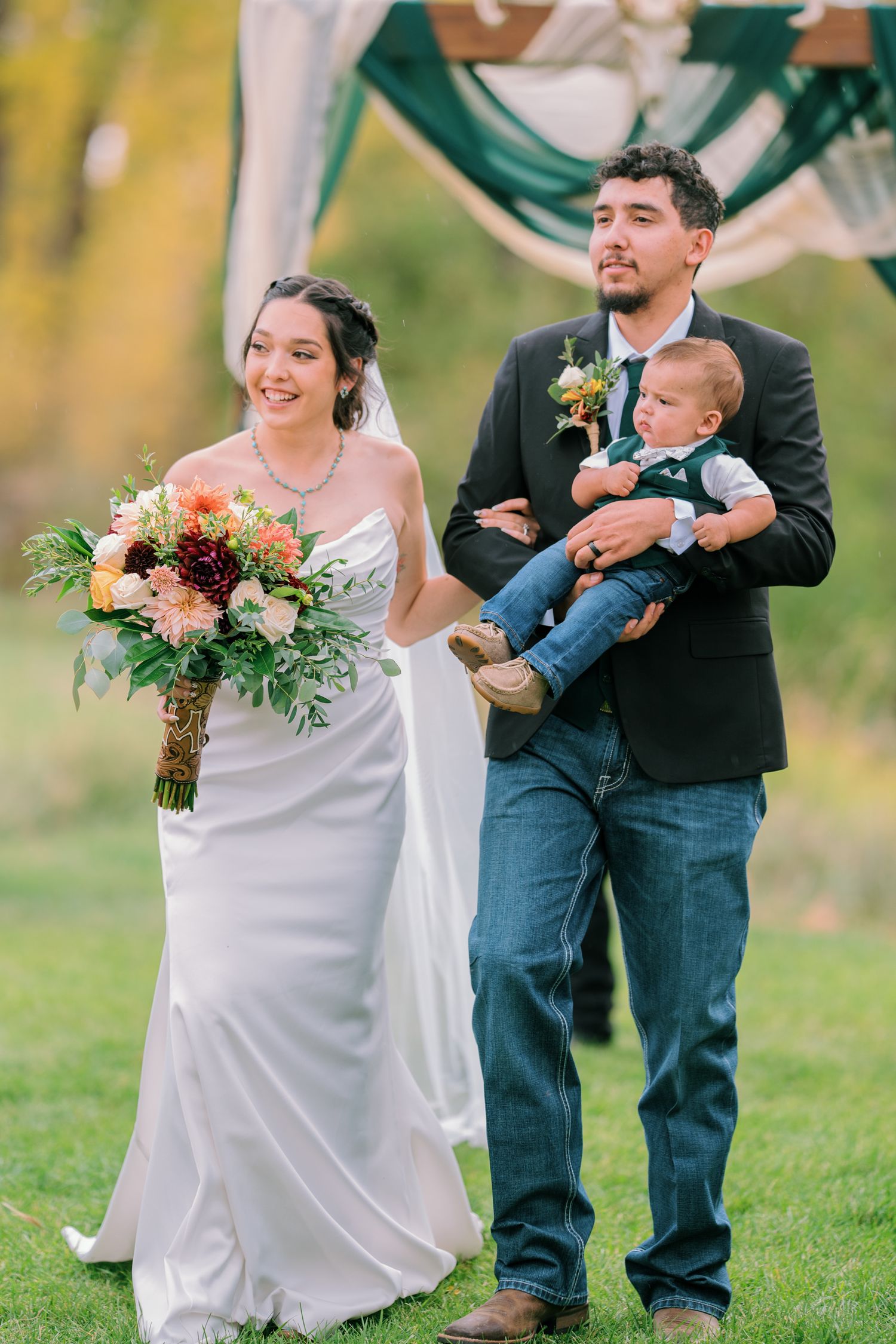 Wedding party with a bride in white dress, groom in casual attire, and baby in outdoor garden setting.