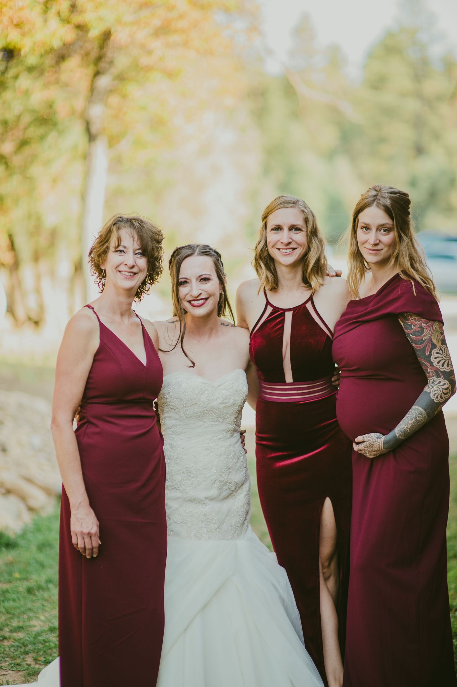 Bridal party in deep burgundy dresses gather for group photo under autumn trees.