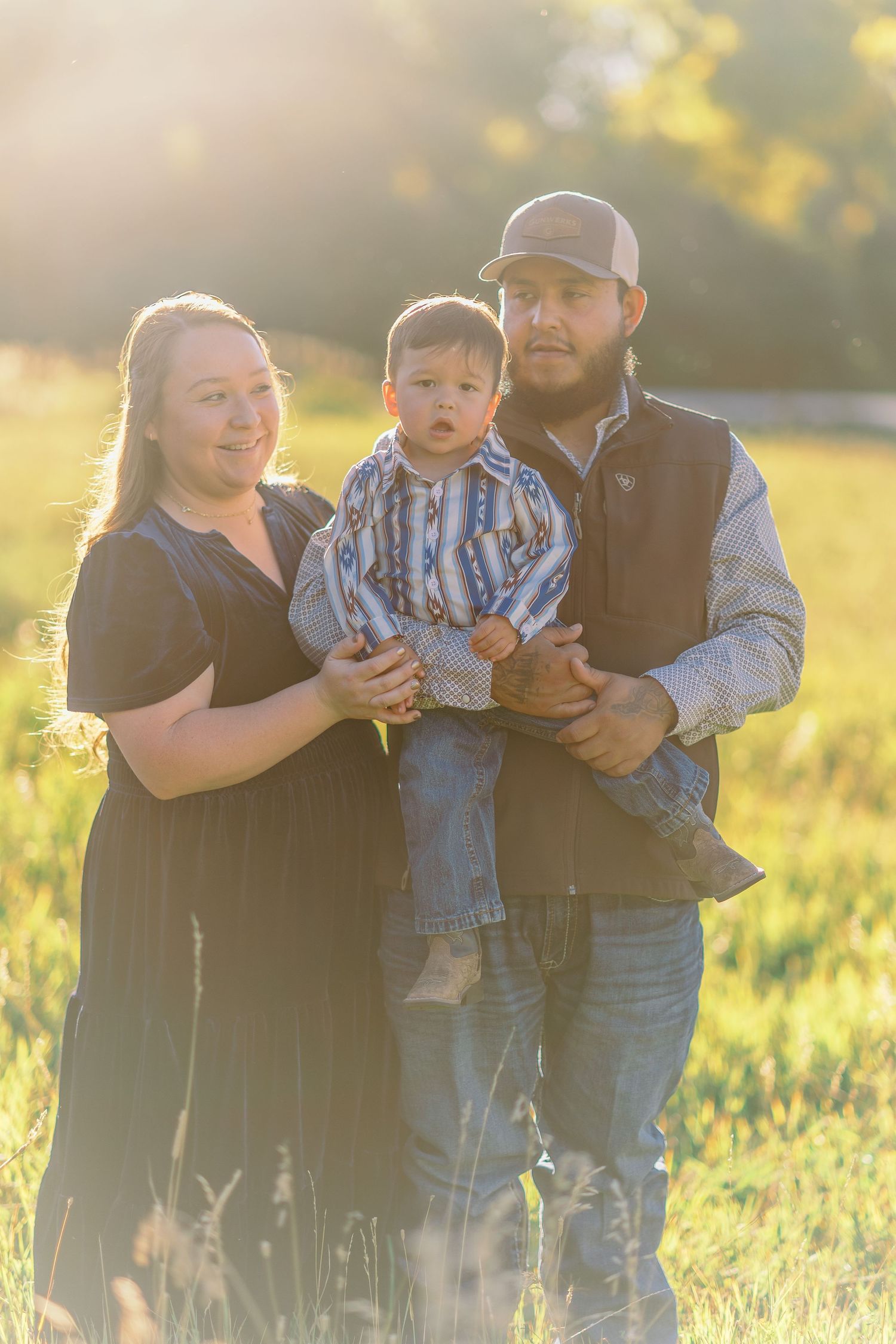 A family poses together in a sunlit meadow at golden hour with warm backlighting and tall grass surrounding them.