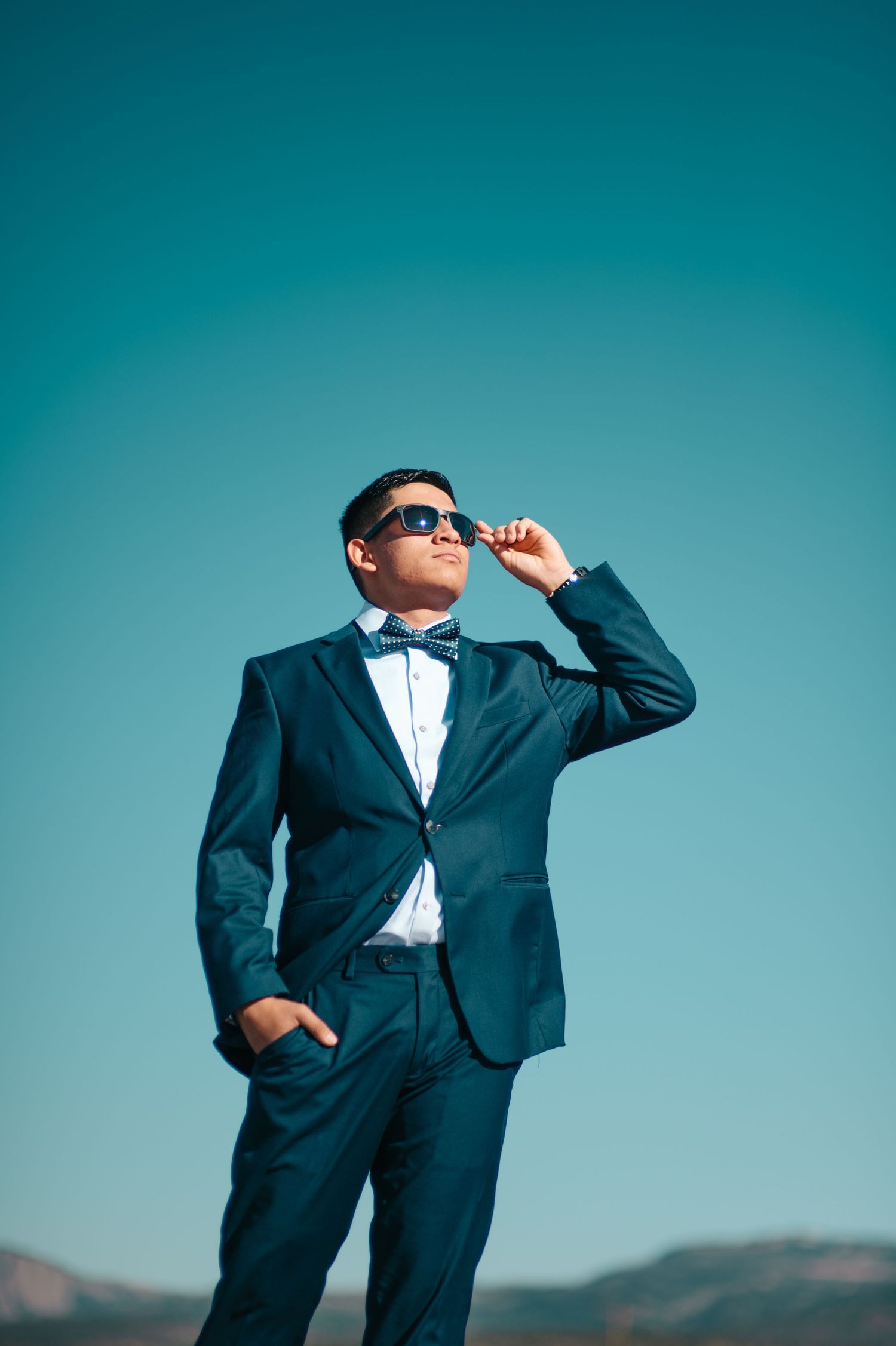 A stylish man in a navy blue tuxedo and bow tie adjusts his sunglasses against a clear blue sky.