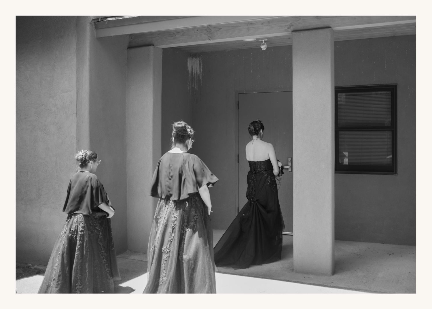 Three women in Victorian-era gowns stand near a doorway in a black and white architectural photograph.