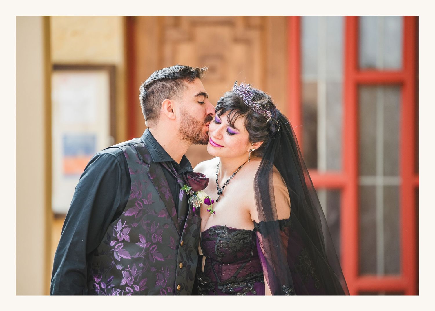 Couple in gothic wedding attire, groom kissing bride's cheek, wearing purple brocade vest and black corset dress.