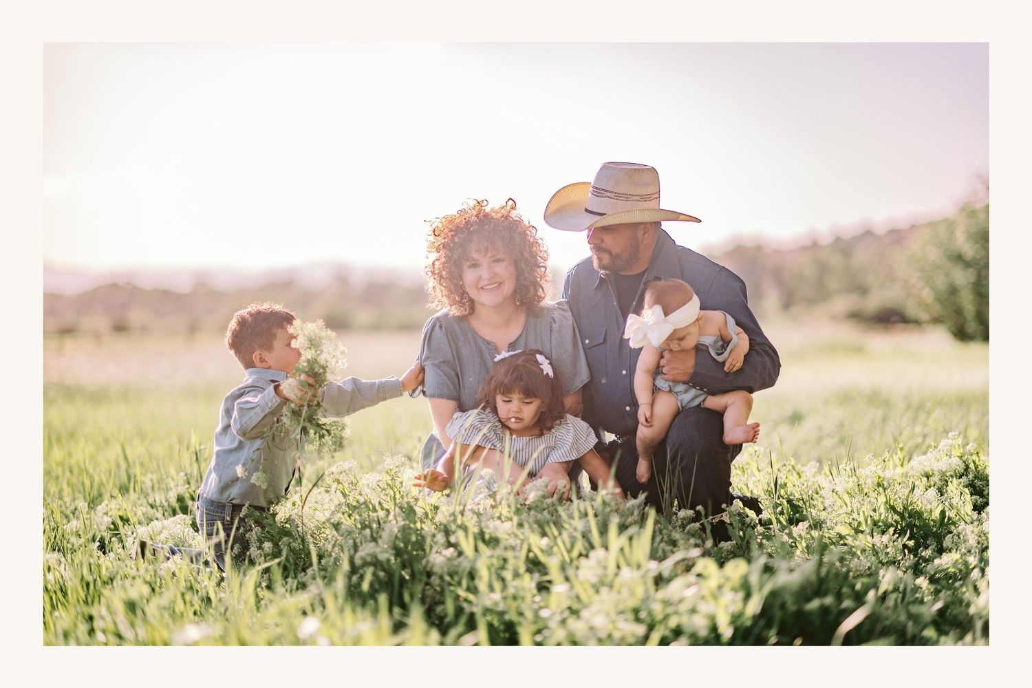 Family of four with two young children and a puppy posing in a sunlit green field for a warm outdoor portrait.