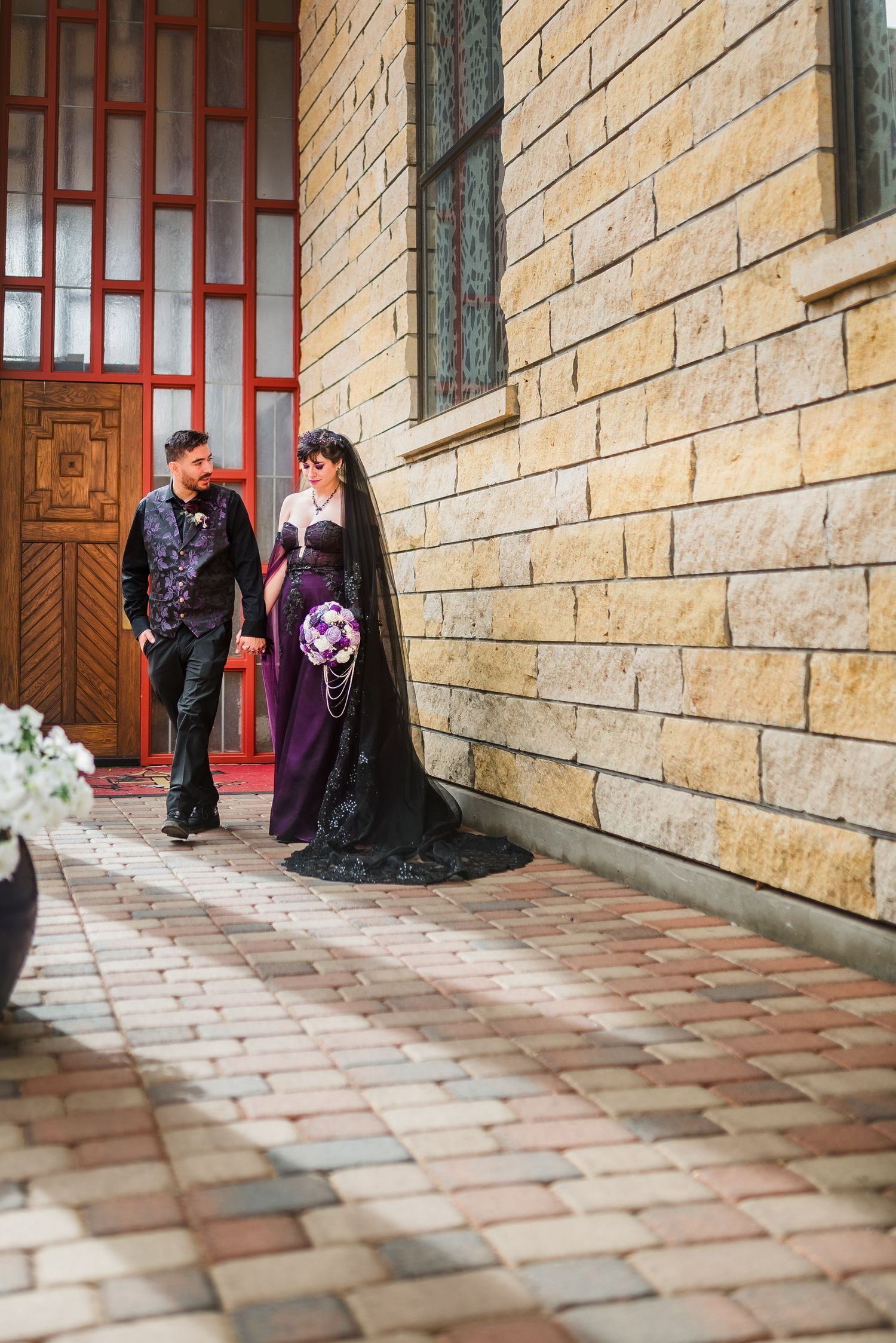 Gothic wedding couple in black and purple attire walking along a stone building with a bouquet.