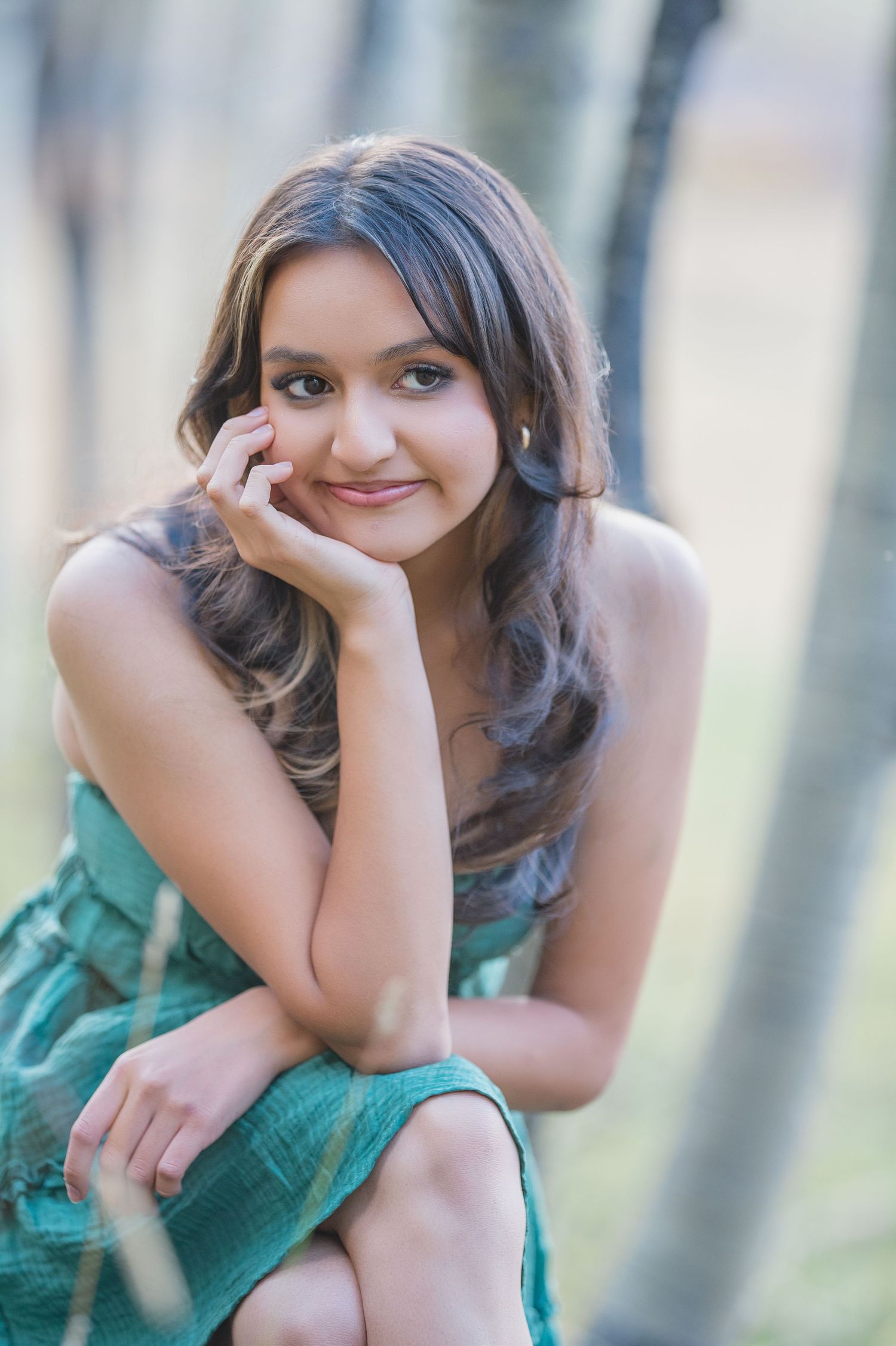 A person in a green dress poses thoughtfully in a soft-focus outdoor setting with blurred trees in the background.