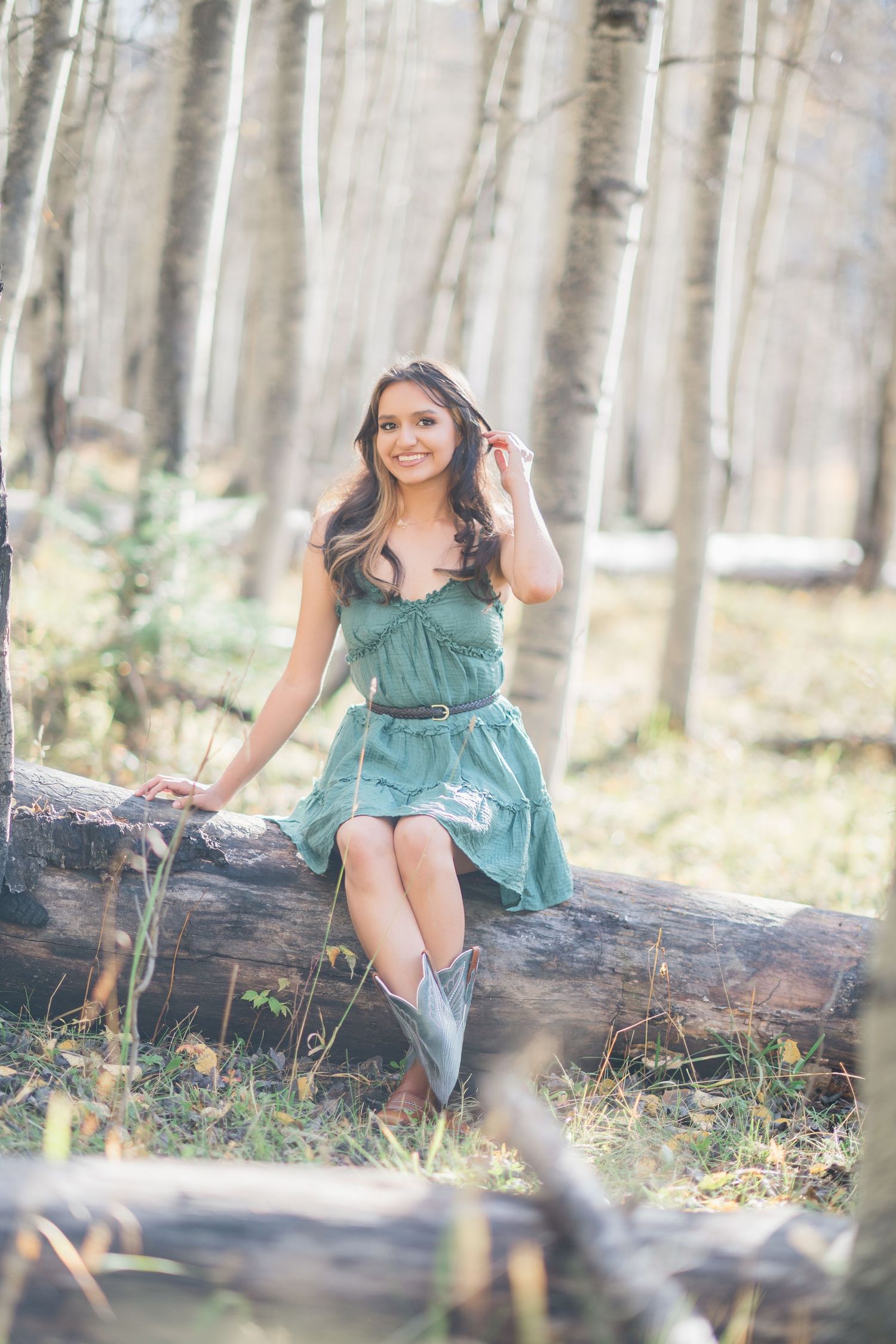 A person in a teal dress and boots sits on a fallen log in a dreamy aspen forest setting.