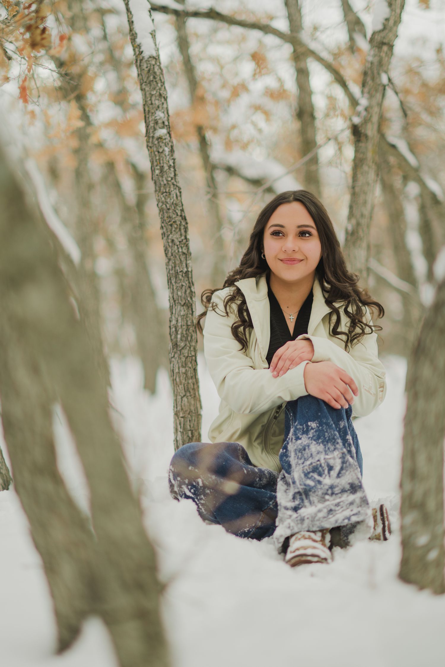 A young woman in winter attire sits in snow-covered woods during a winter portrait photography session.