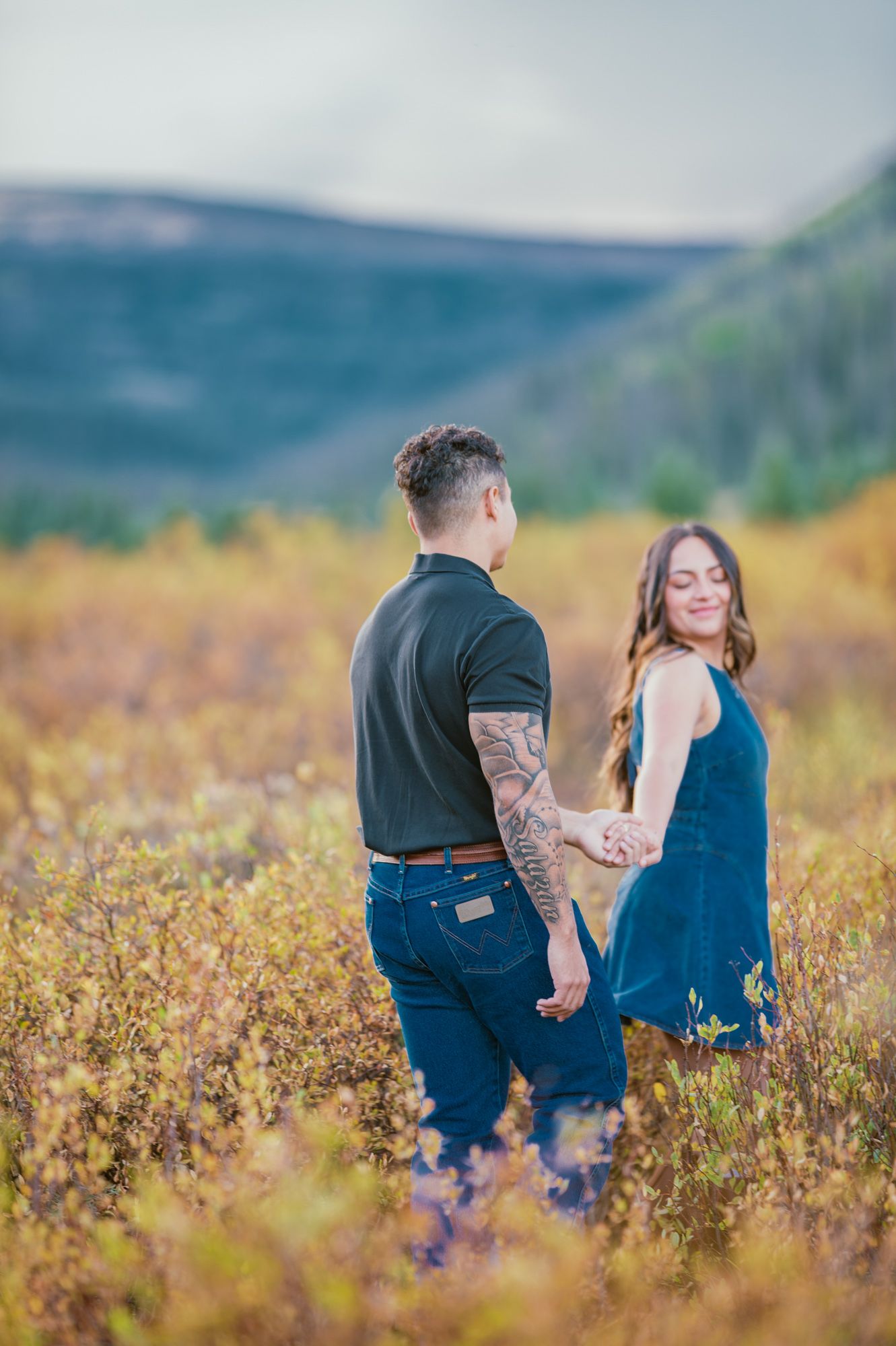 Couple holding hands in a golden field with mountain backdrop, man with tattoos in navy polo and woman in teal dress.