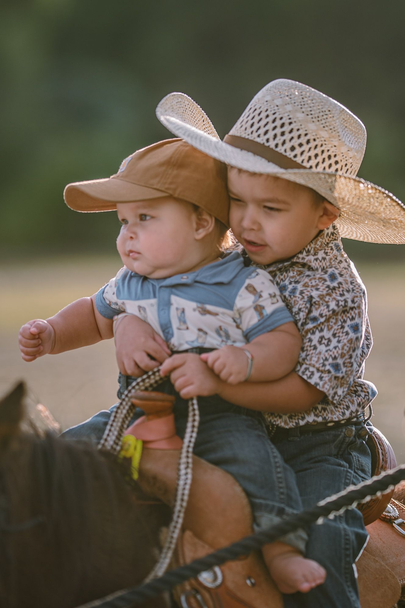 Two toddlers in cowboy hats sit together on a horse, dressed in western outfits in a sunny outdoor setting.