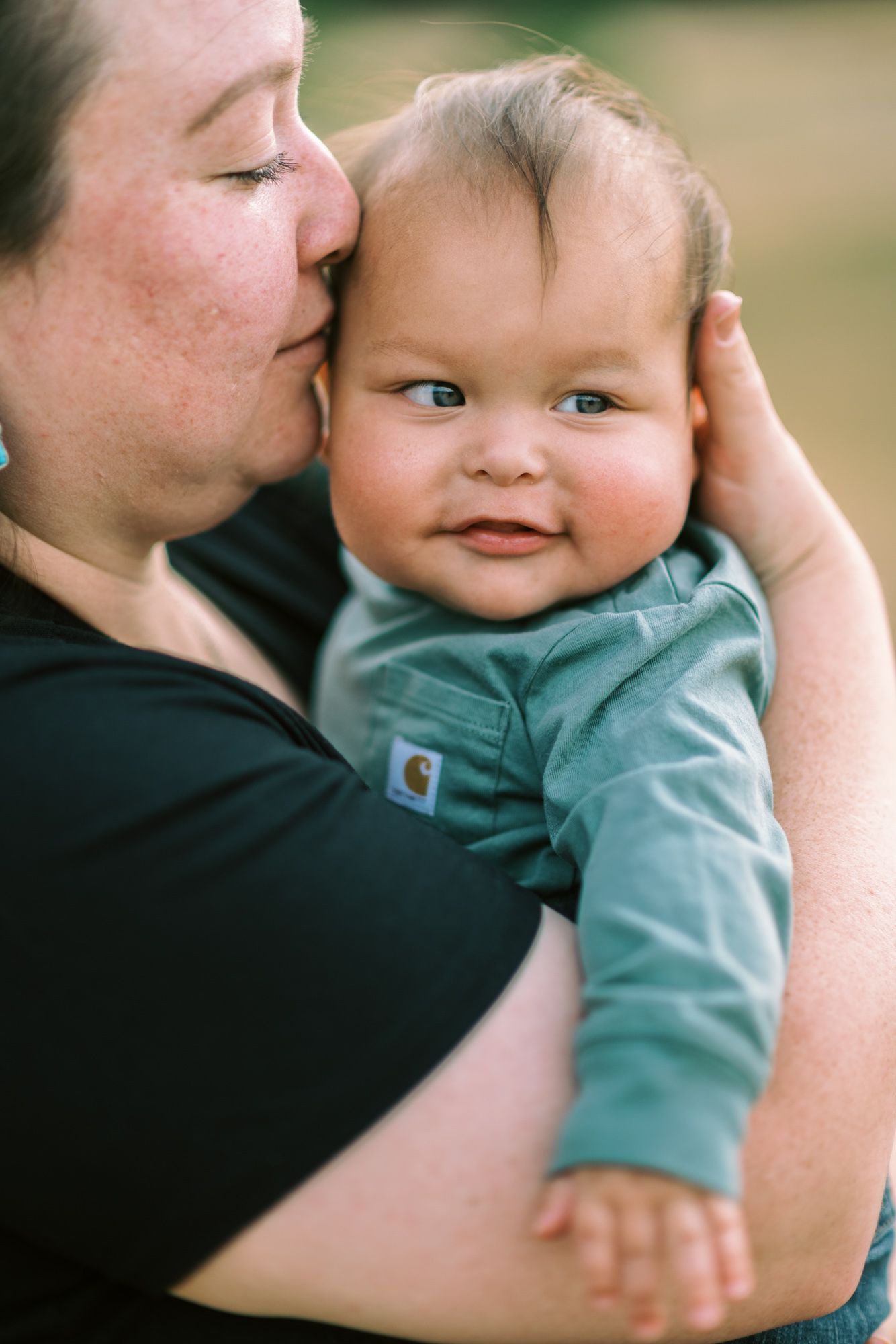A mother tenderly kisses her smiling baby boy dressed in a green Carhartt onesie outdoors.