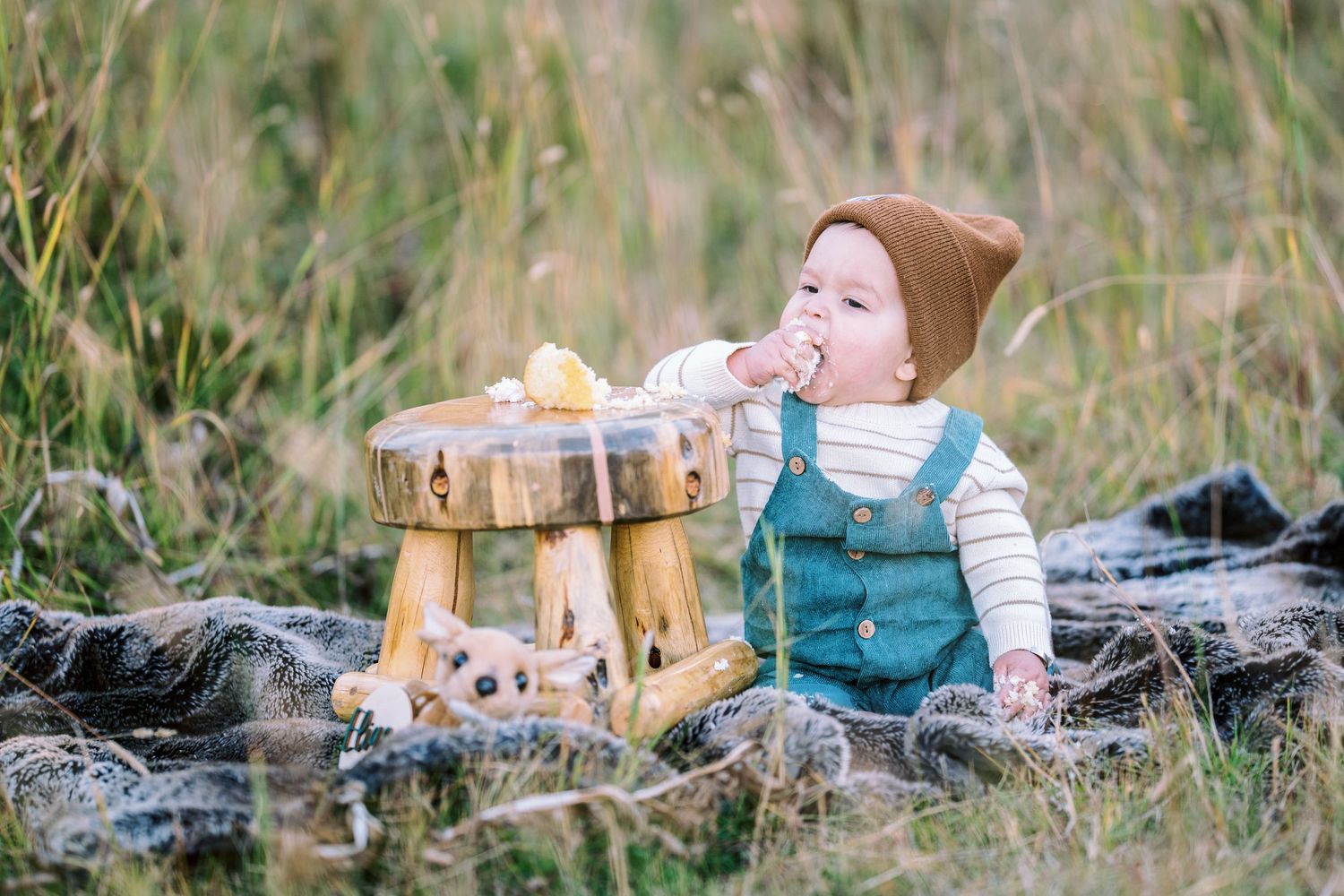 A baby in teal overalls sits beside a wooden stool in tall grass.