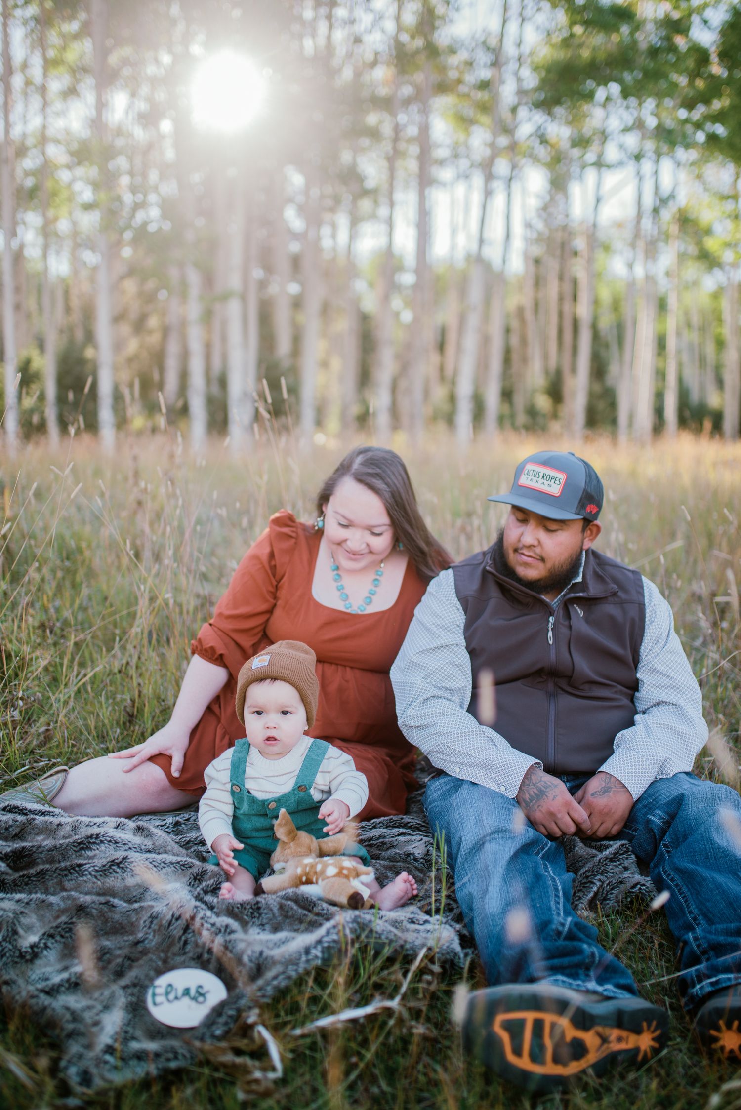 Family sitting together on blanket in sunlit forest clearing during portrait session.