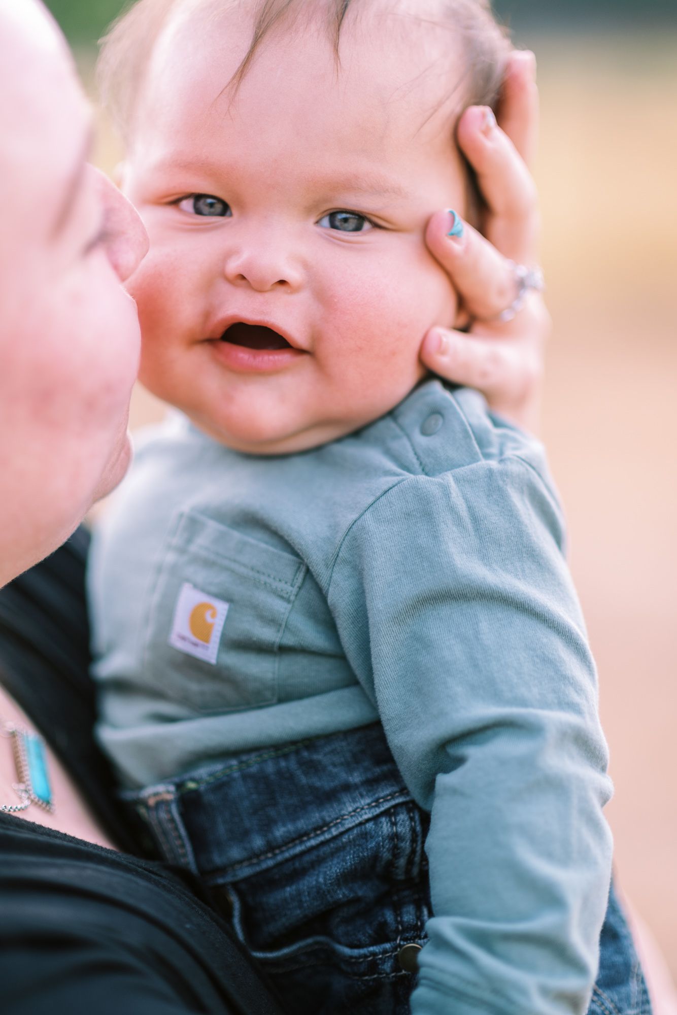 Smiling baby in Carhartt outfit being held by mother outdoors, showing chubby cheeks and bright blue eyes.
