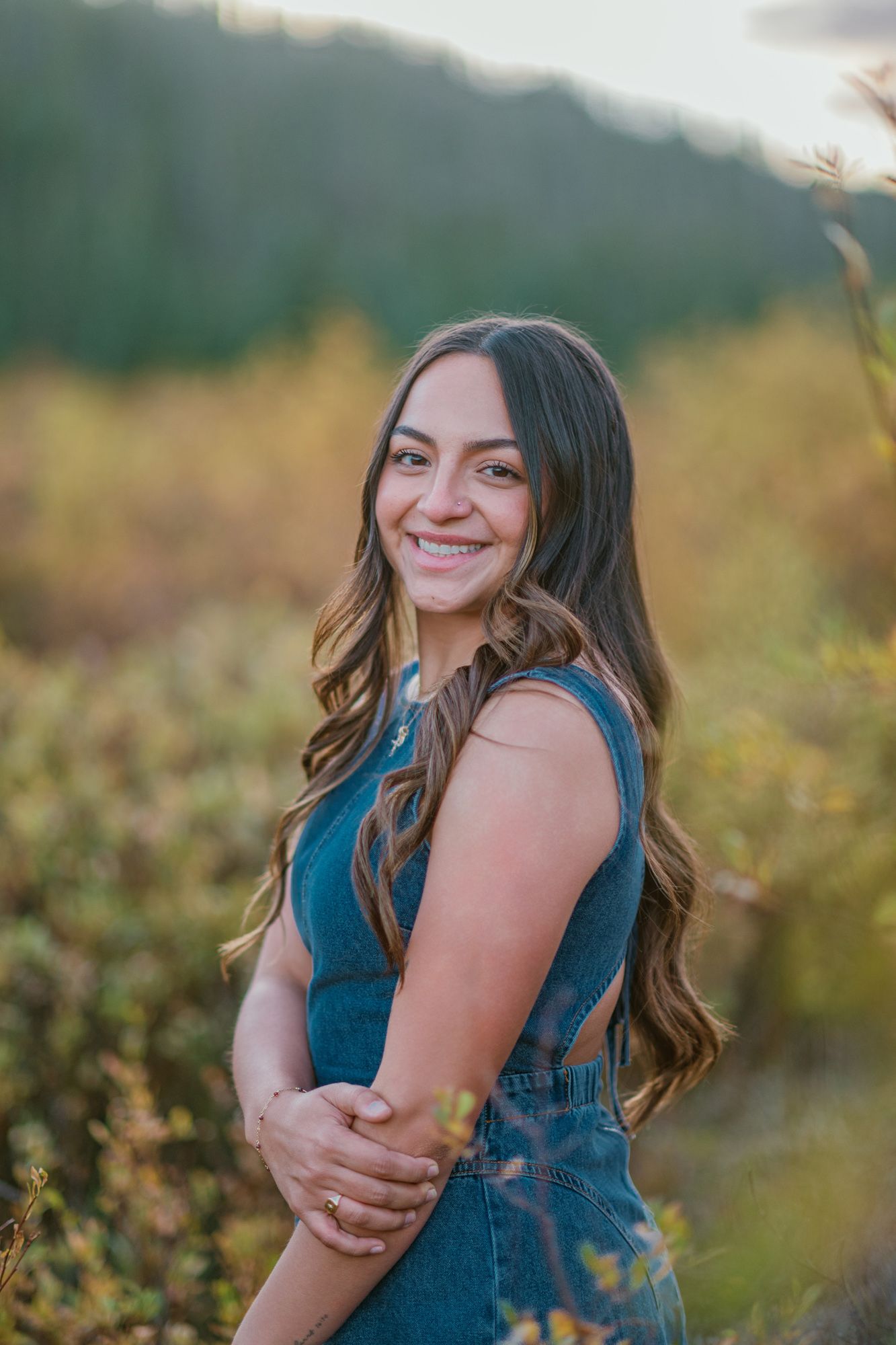 Young woman with long wavy hair smiling outdoors in denim overalls, surrounded by autumn foliage and green hills.