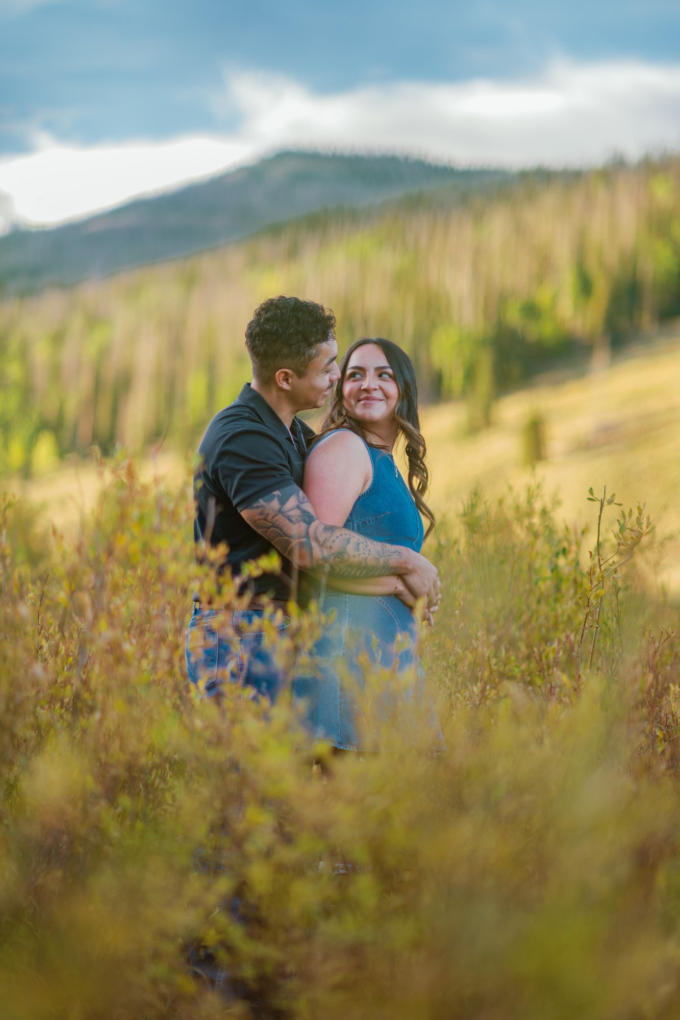 Couple embracing in a golden meadow with mountain backdrop, man kissing woman's cheek during outdoor engagement session.
