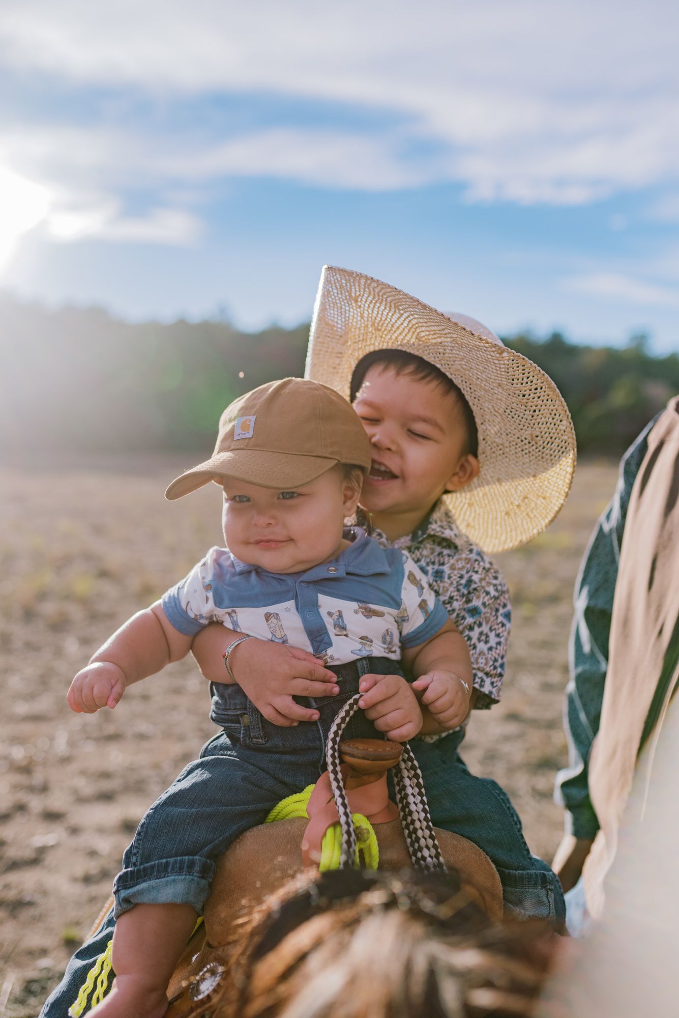 Two young children in cowboy hats sitting on a horse outdoors under a bright blue sky.