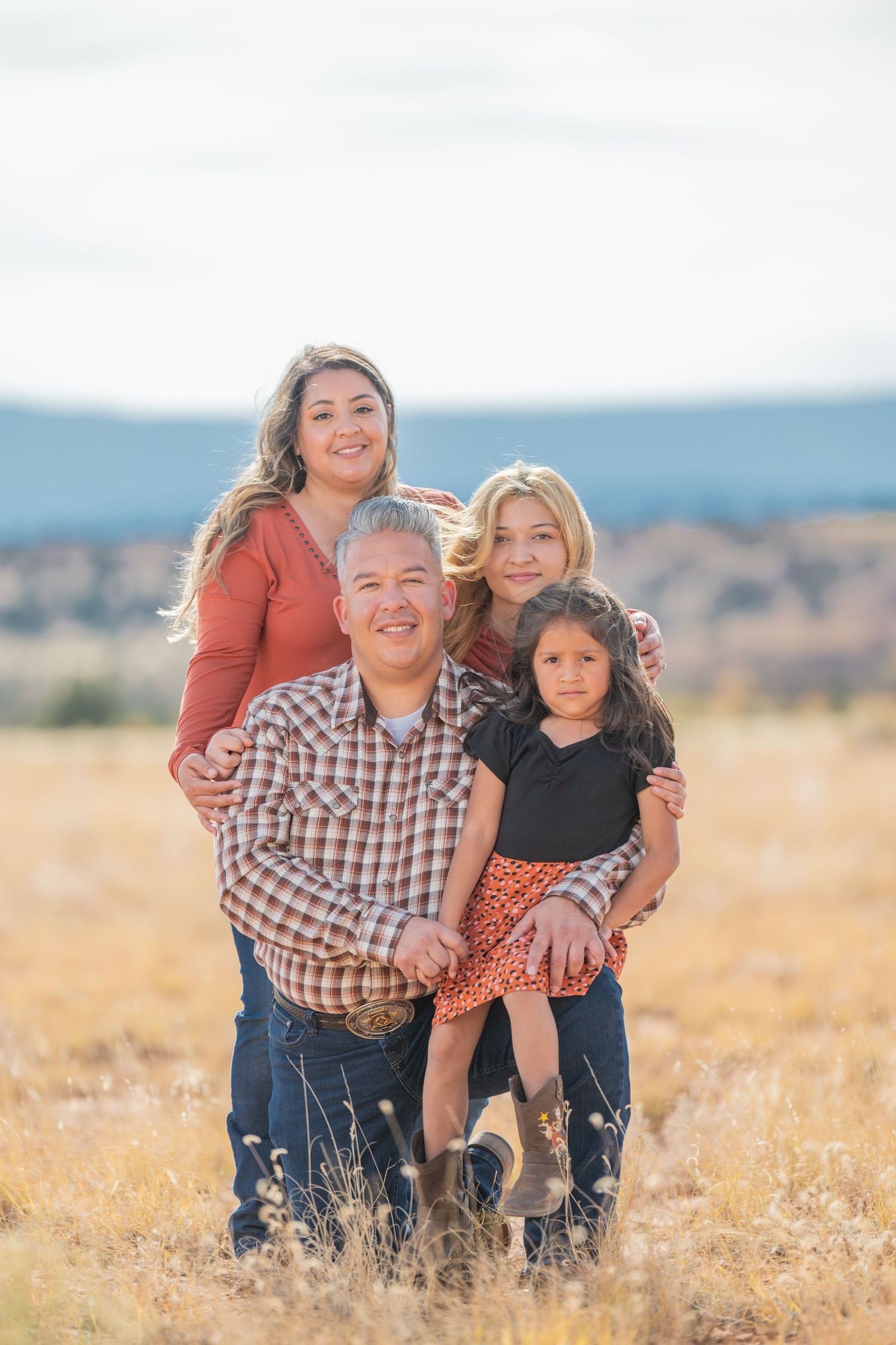 Three people pose together in a golden field during sunset with mountains visible in the background.