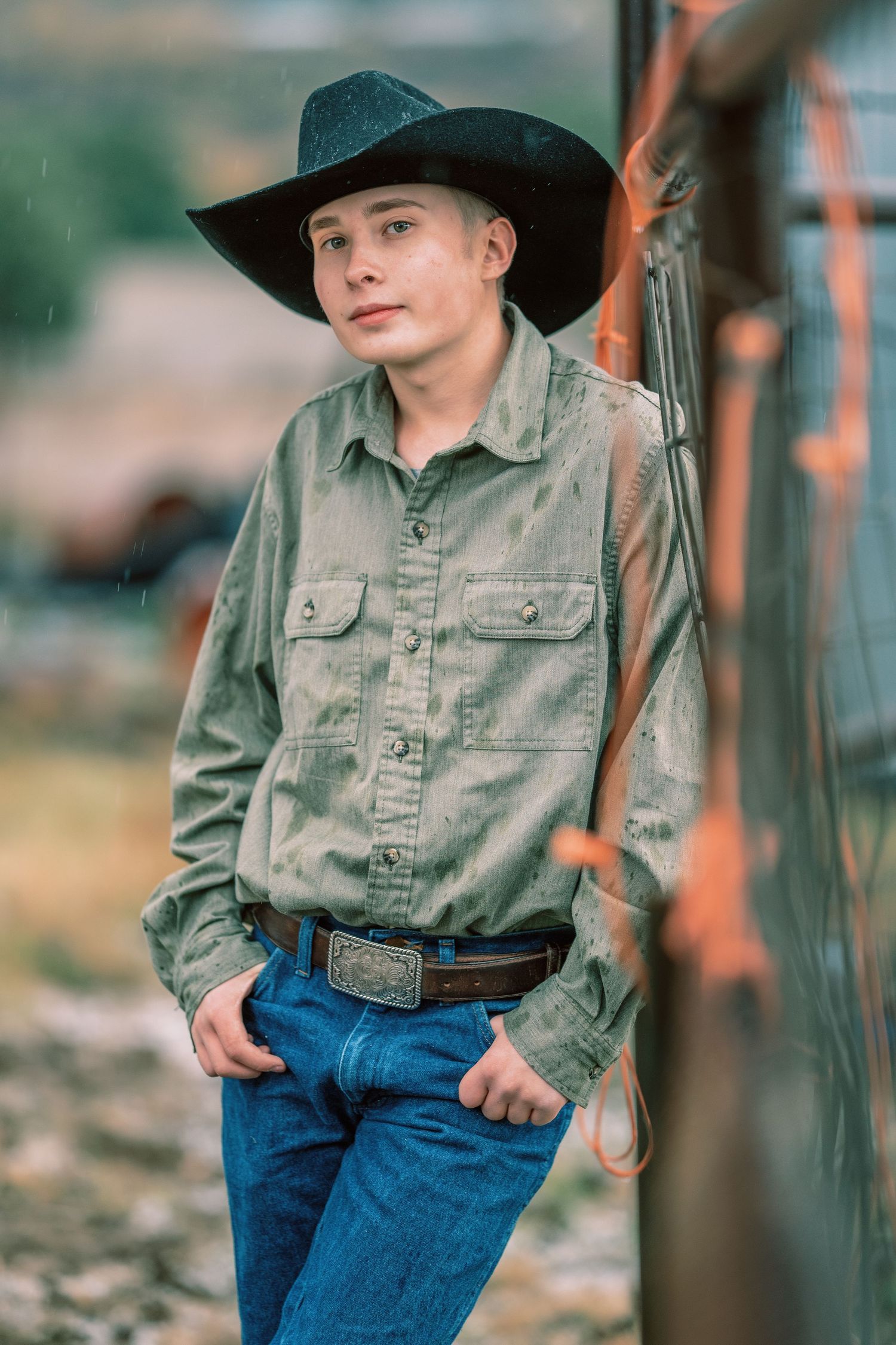 Young rancher wearing black cowboy hat, olive shirt and jeans stands confidently in western attire outdoors.