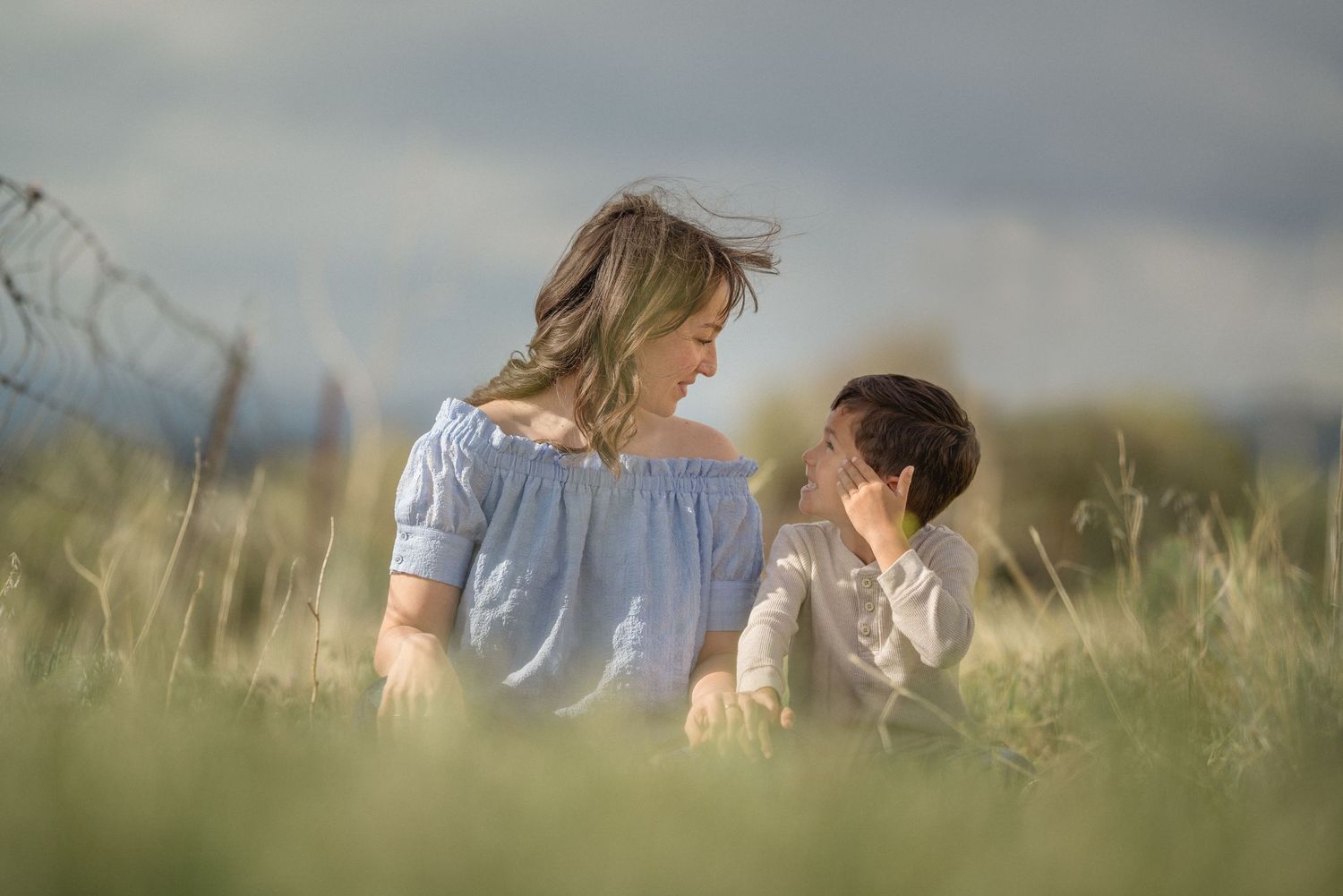Two people sit close together in a peaceful field of tall grass during golden hour as wind gently blows.