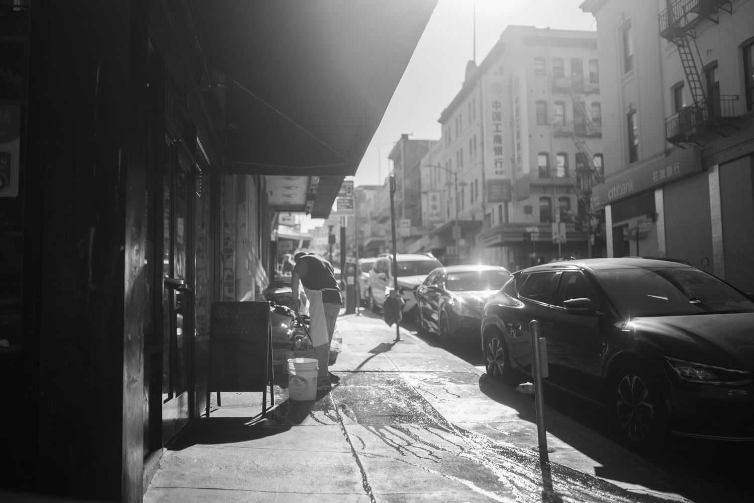 Black and white urban street scene on a sunny day with strong diagonal shadows across parked cars and buildings.