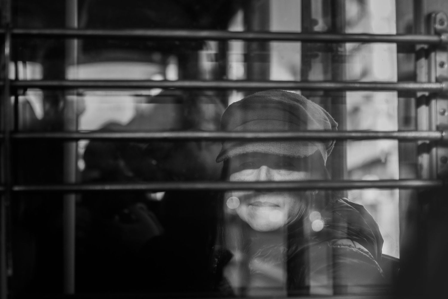 A person smiles while wearing sunglasses and sitting in front of bright window blinds in black and white.