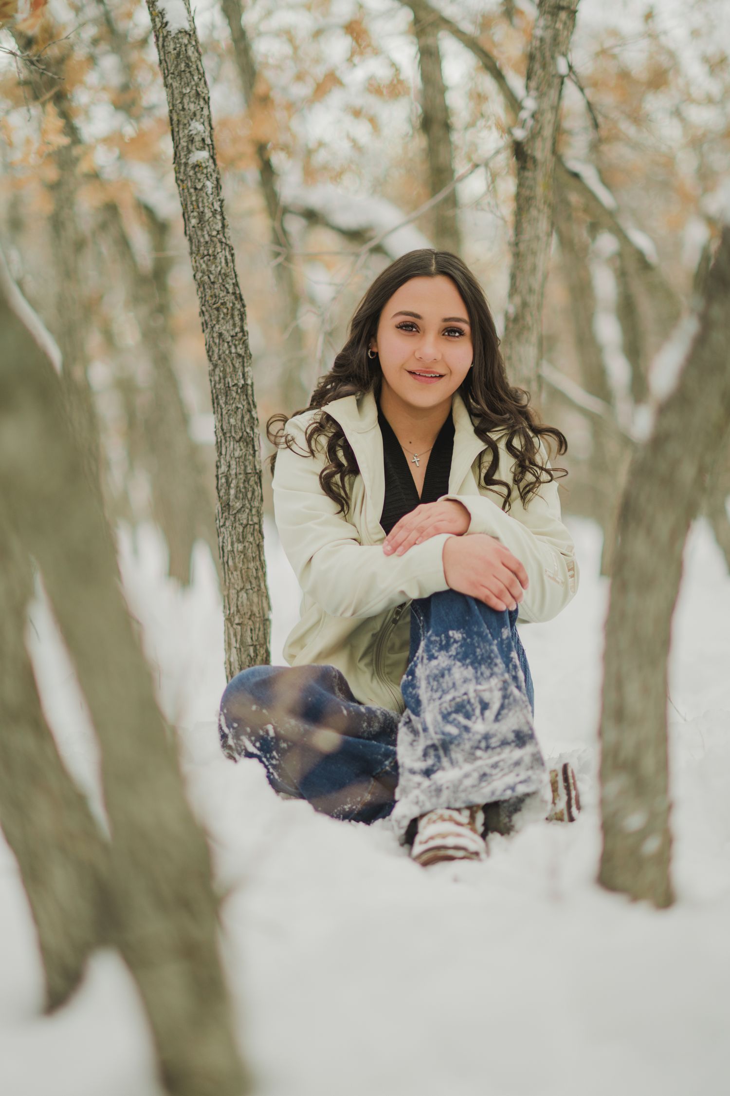 Winter portrait in snowy woods with person wearing white coat and jeans sitting among birch trees.