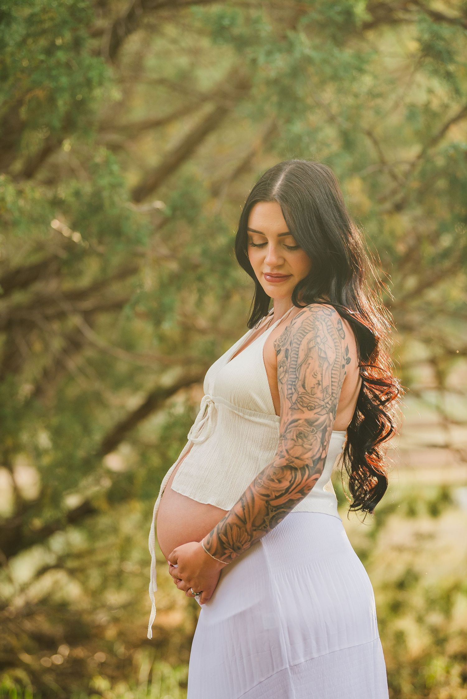 Pregnant woman with sleeve tattoos in white dress holding baby bump outdoors among green trees.