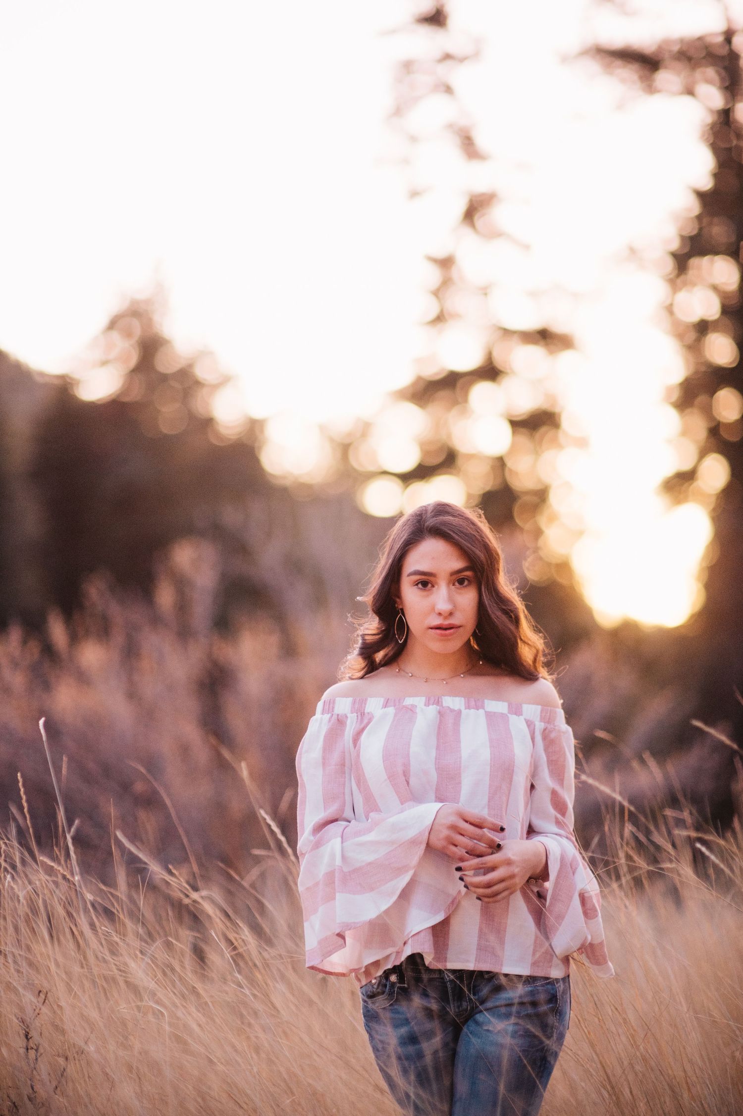 Young woman in off-shoulder striped top and jeans standing in golden field at sunset.