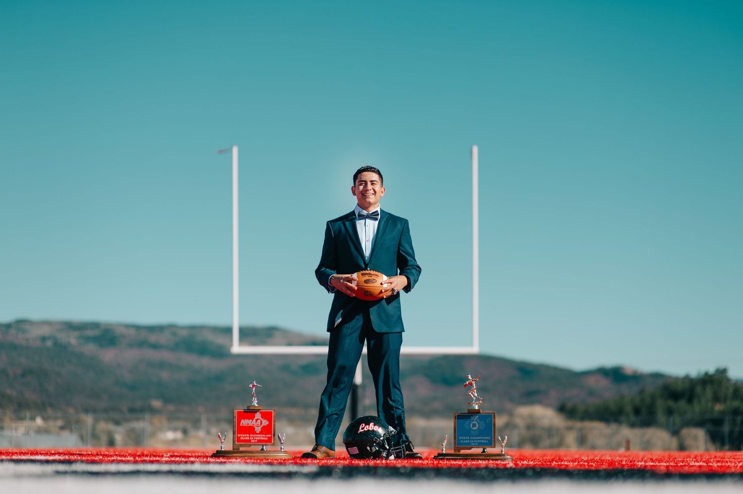 Young man in teal suit holding football on field with goalposts and mountain backdrop.