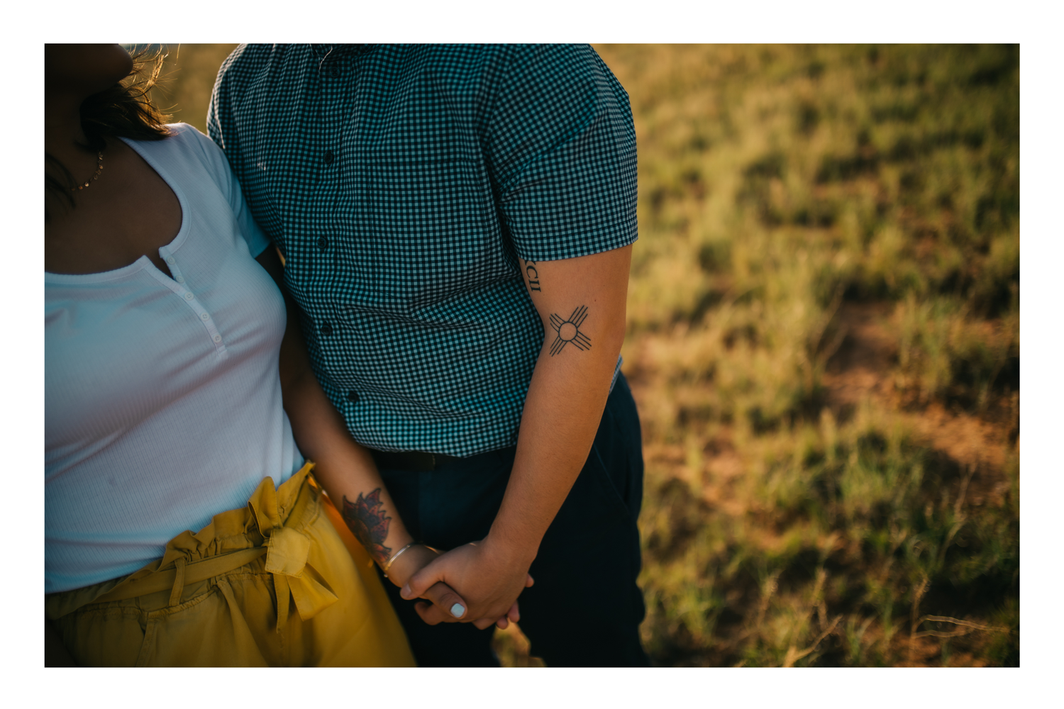 Couple holding hands in a golden field, woman in white top and yellow skirt, man with tattoo in teal shirt.