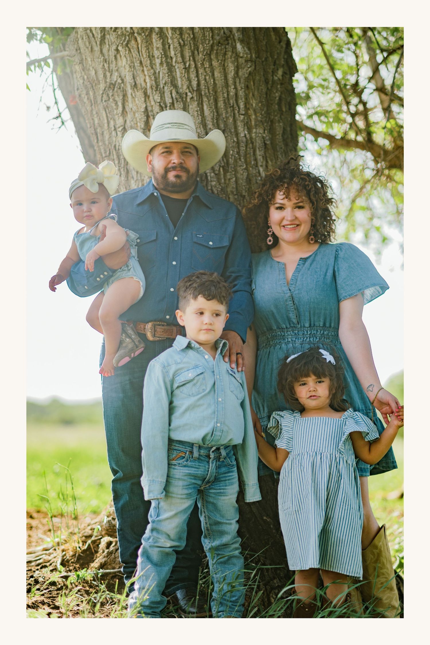 Family of five posing outdoors by a tree, wearing coordinated blue outfits, with father in cowboy hat holding baby girl.