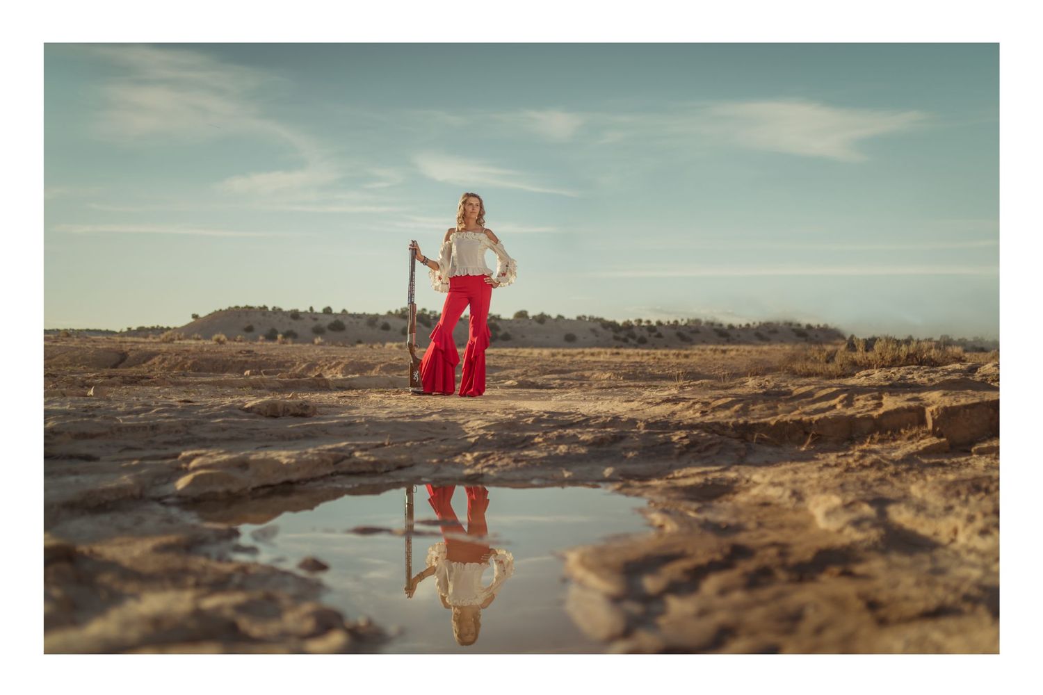 Woman in red flared pants and white top stands on rocky terrain with her reflection visible in a puddle below.