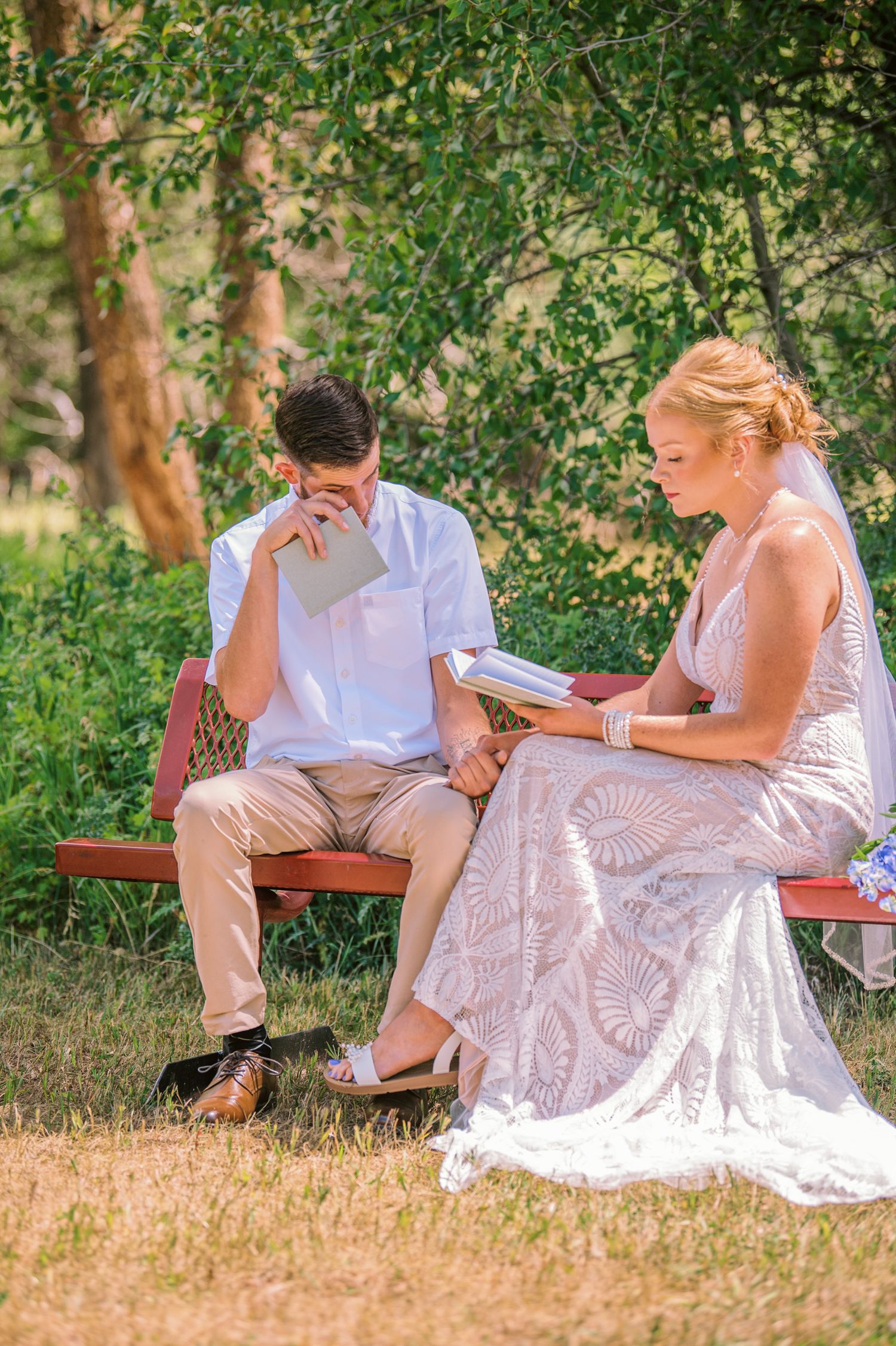 Bride and groom sitting on a bench reading vows outdoors on their wedding day surrounded by lush green trees.