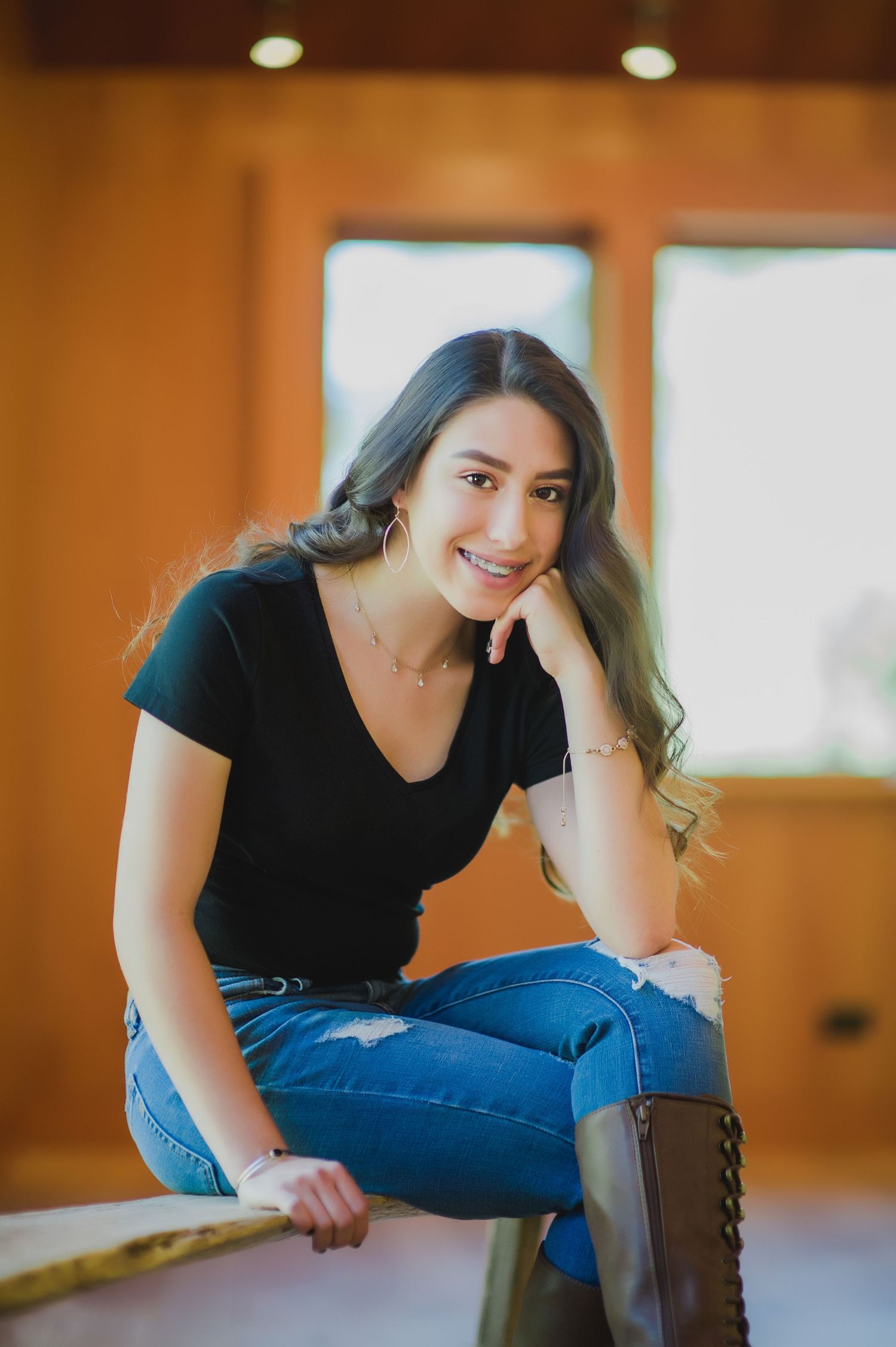 Young woman in black t-shirt and jeans smiling while sitting relaxed in a warmly lit wooden room.