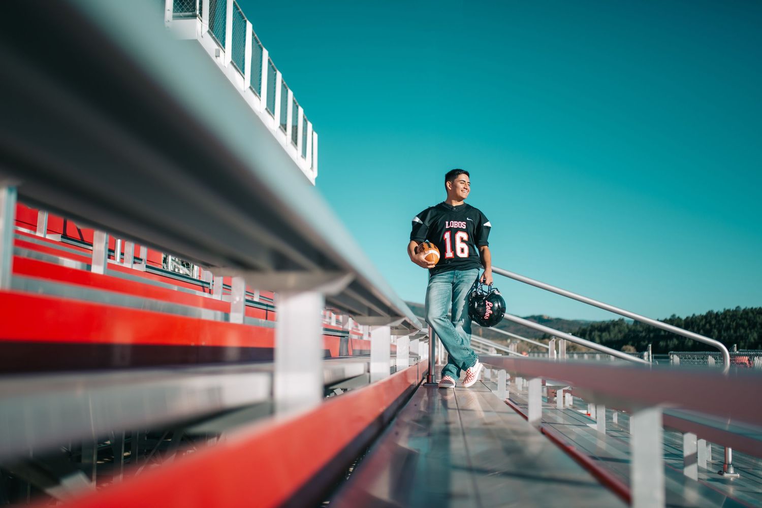 Football player wearing jersey number 16 stands in stadium bleachers holding helmet on a sunny day.