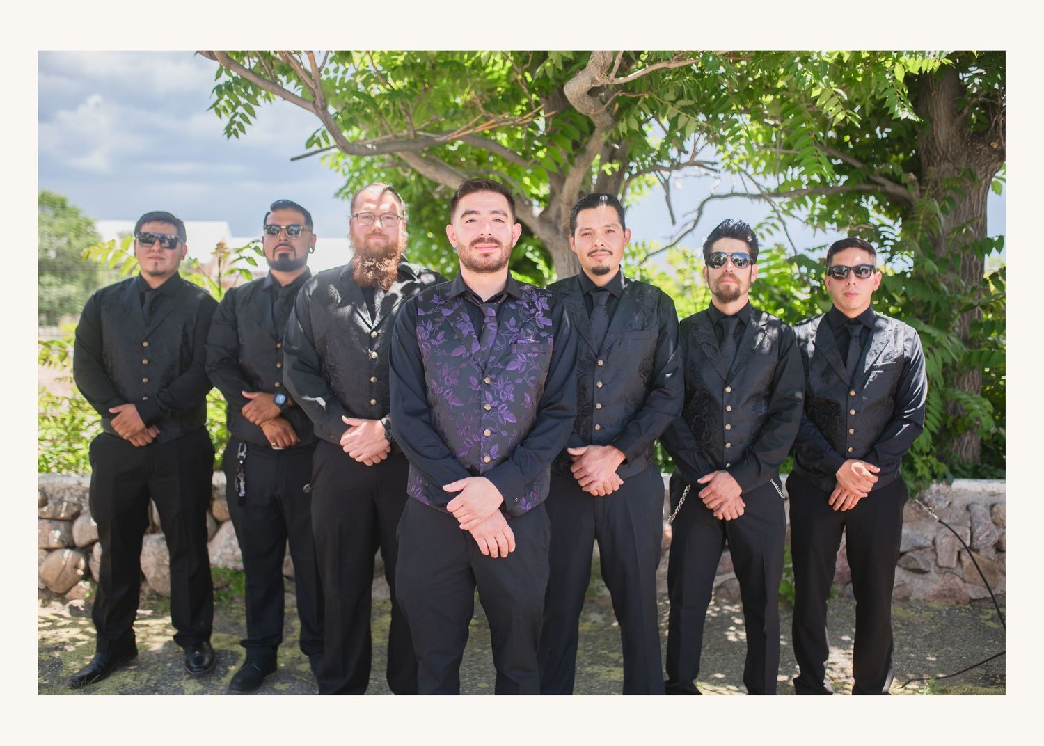 Groom and groomsmen in black suits pose outdoors under a tree at a tropical wedding venue.
