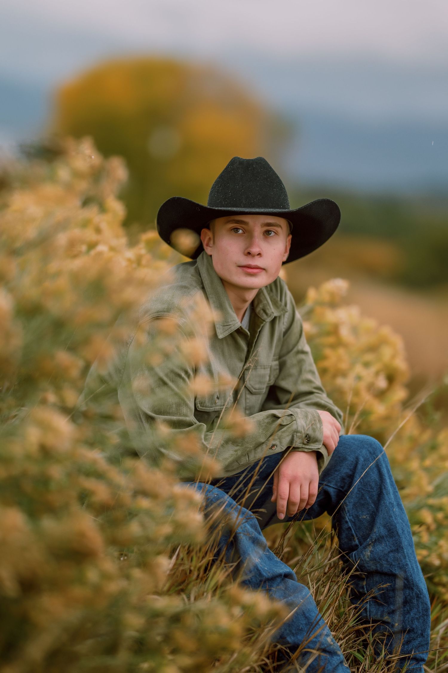 A person in a black cowboy hat and denim sits in tall golden grass against a soft natural background.