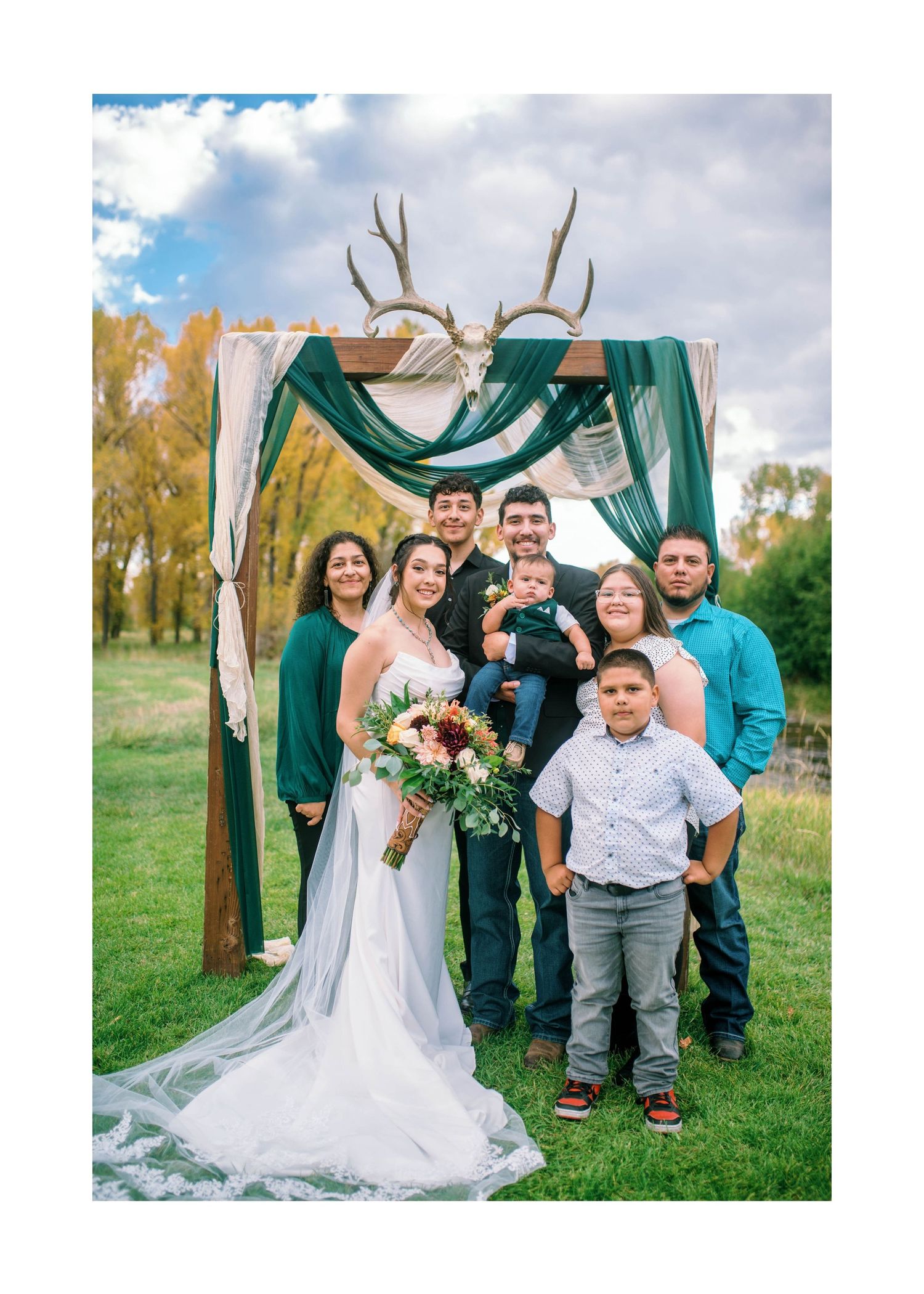 A wedding party gathers under a draped green arch with antlers outdoors in front of autumn trees.