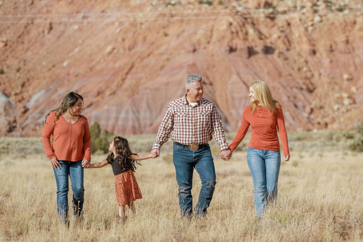 People walk hand in hand through a golden field with red rock formations in the background.